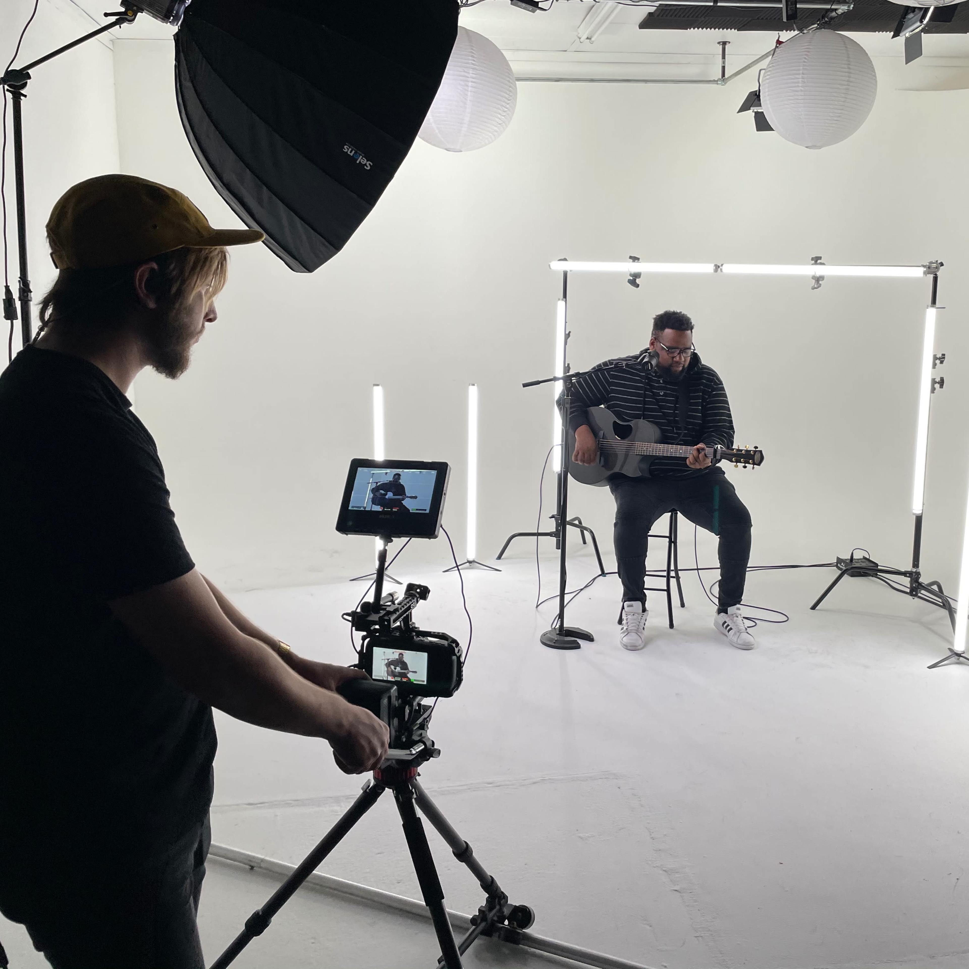 A musician plays guitar on a stool in a brightly lit studio surrounded by lighting equipment and film crew members.