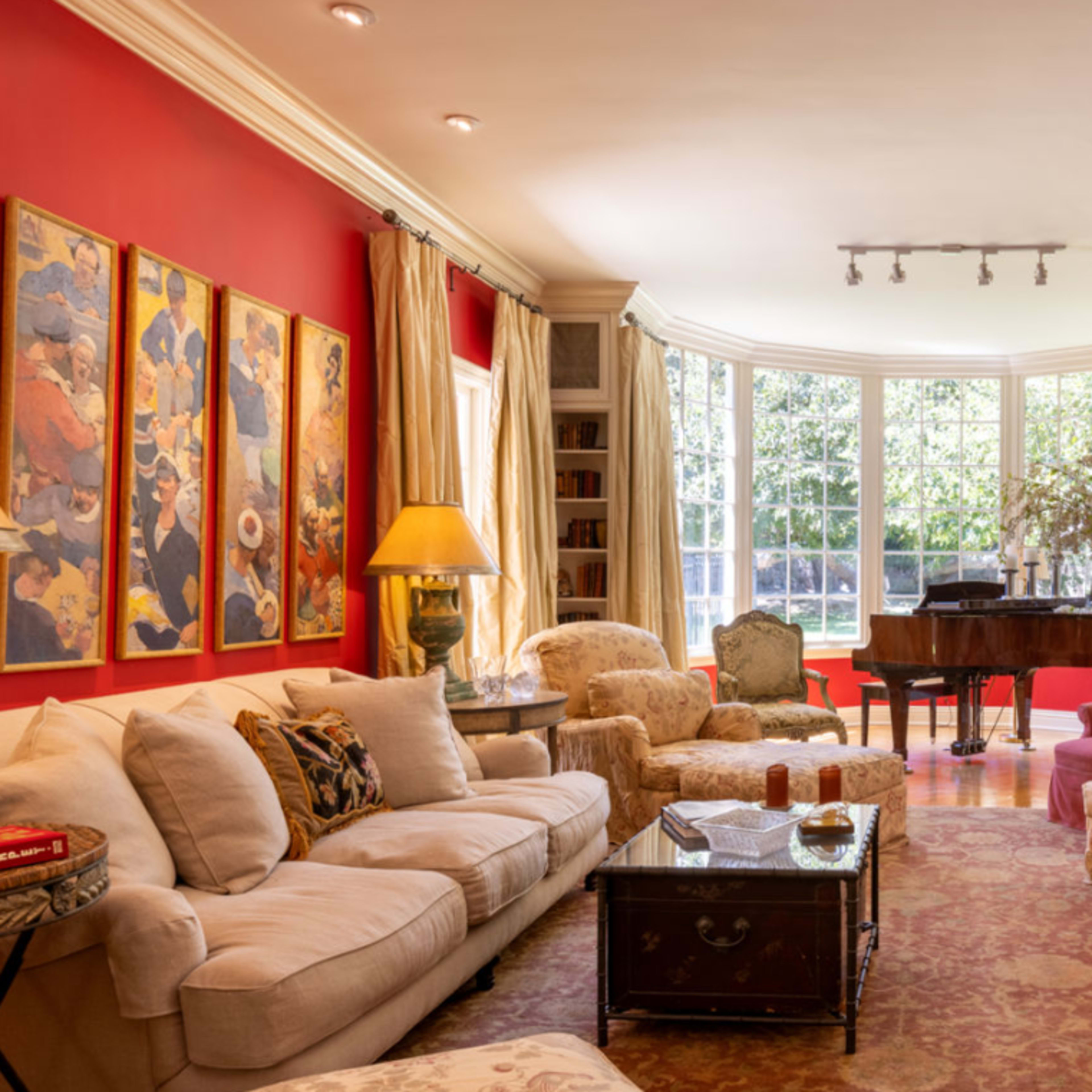 A bright living room with red walls, large windows, a grand piano, and various seating arrangements, including couches and armchairs.