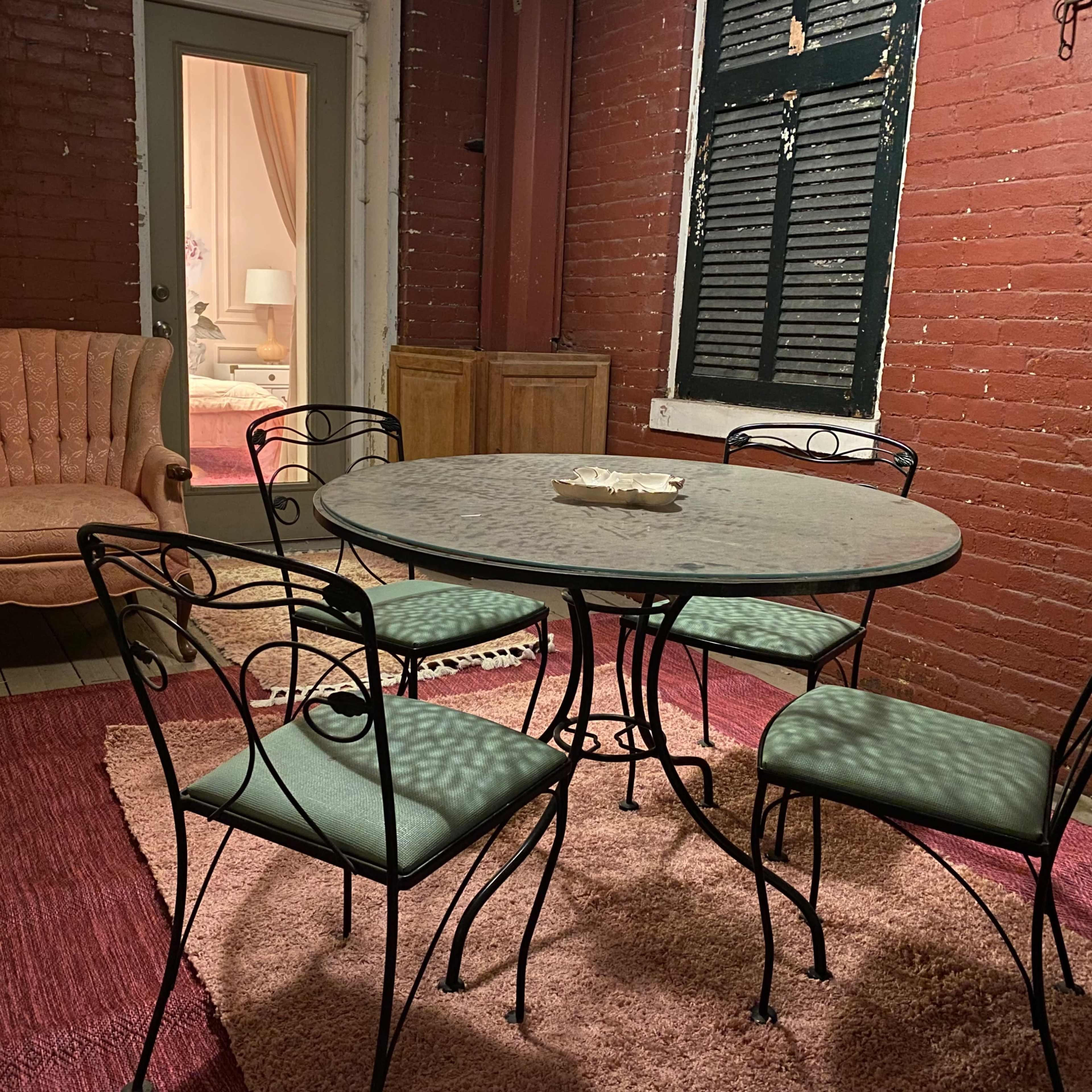 A round metal table with four chairs is set on a rug in a red-brick room, with a pink armchair visible in the doorway.