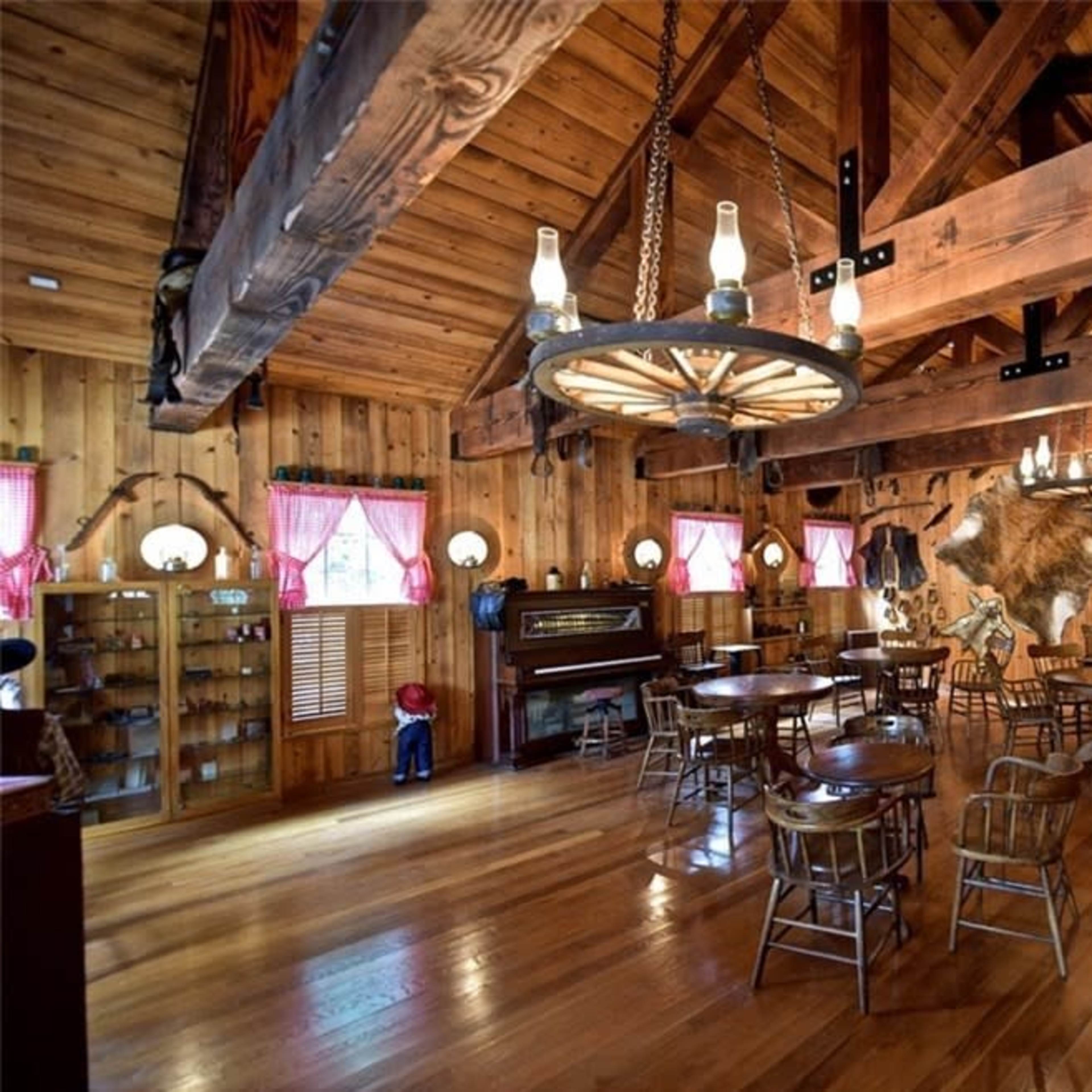 A wooden interior of a rustic dining or gathering space, featuring exposed beams, several round tables, and decorations including a mounted animal head and a piano.