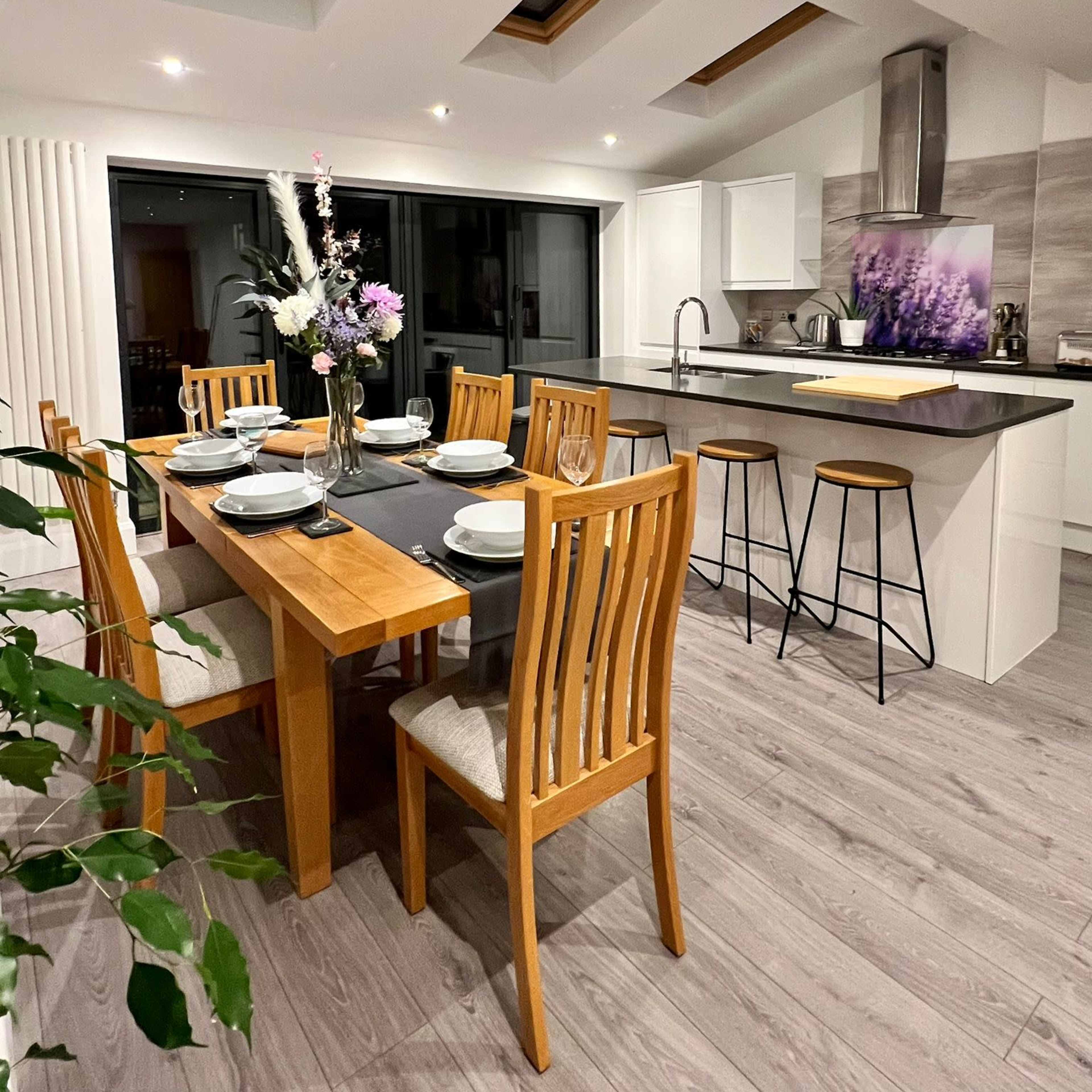 A modern kitchen features a dining table with eight chairs, skylights above, a counter with bar stools, and a floral centerpiece.