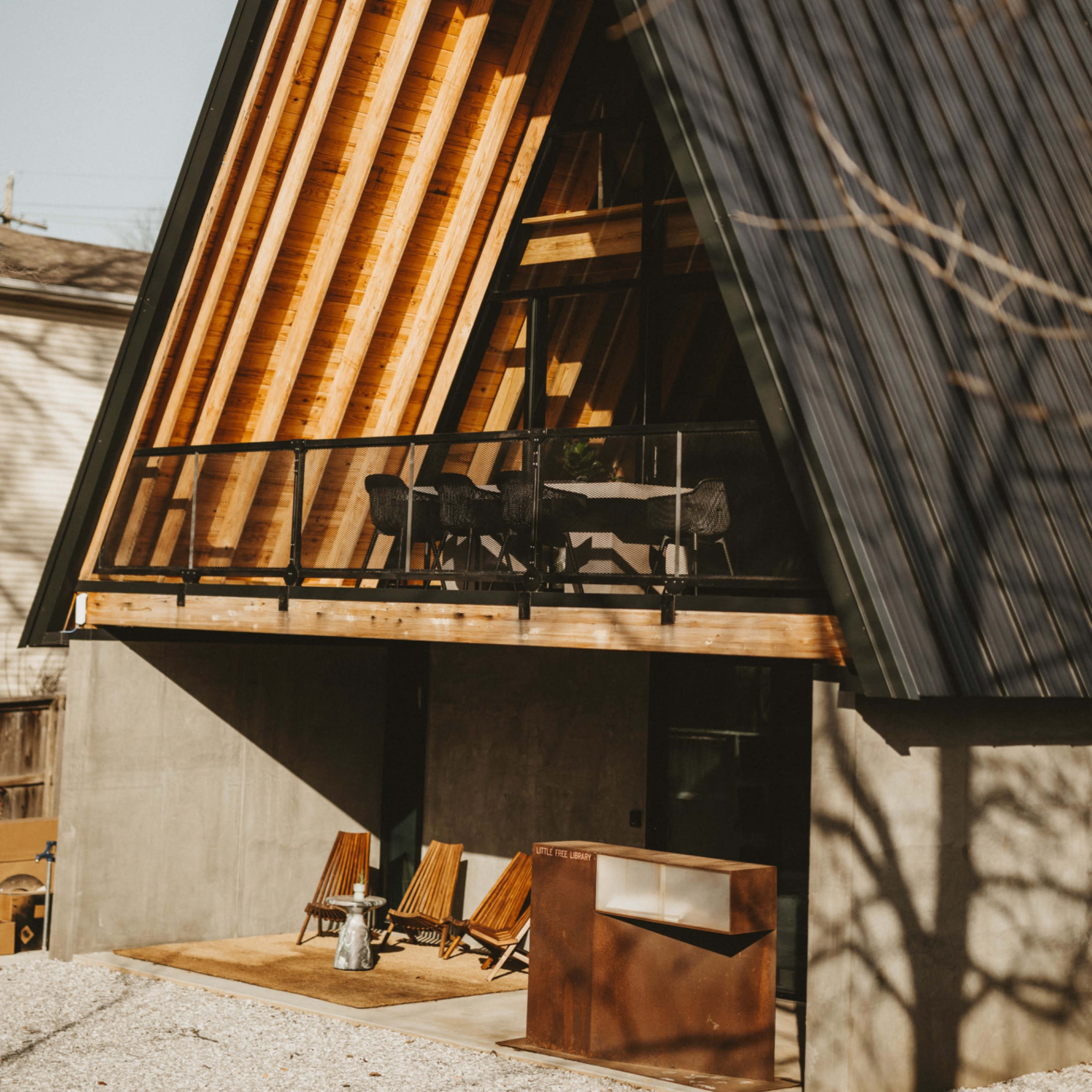 A modern A-frame house with a triangular roof, wooden beams, and a concrete base, featuring a balcony and patio furniture in the foreground.