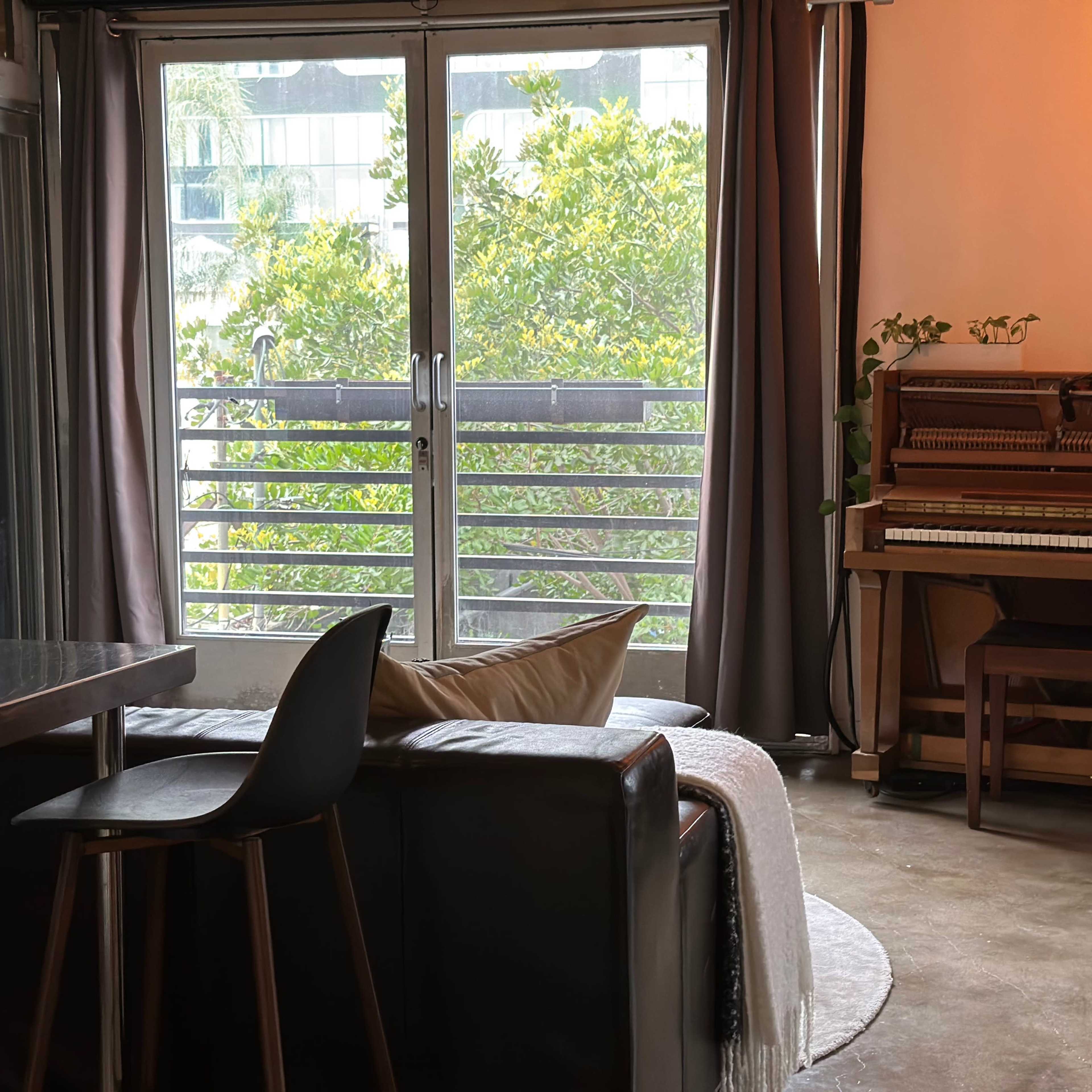 A modern living room with a black sofa, a dining area with a metal table, and a piano next to a large window that offers a view of greenery outside.