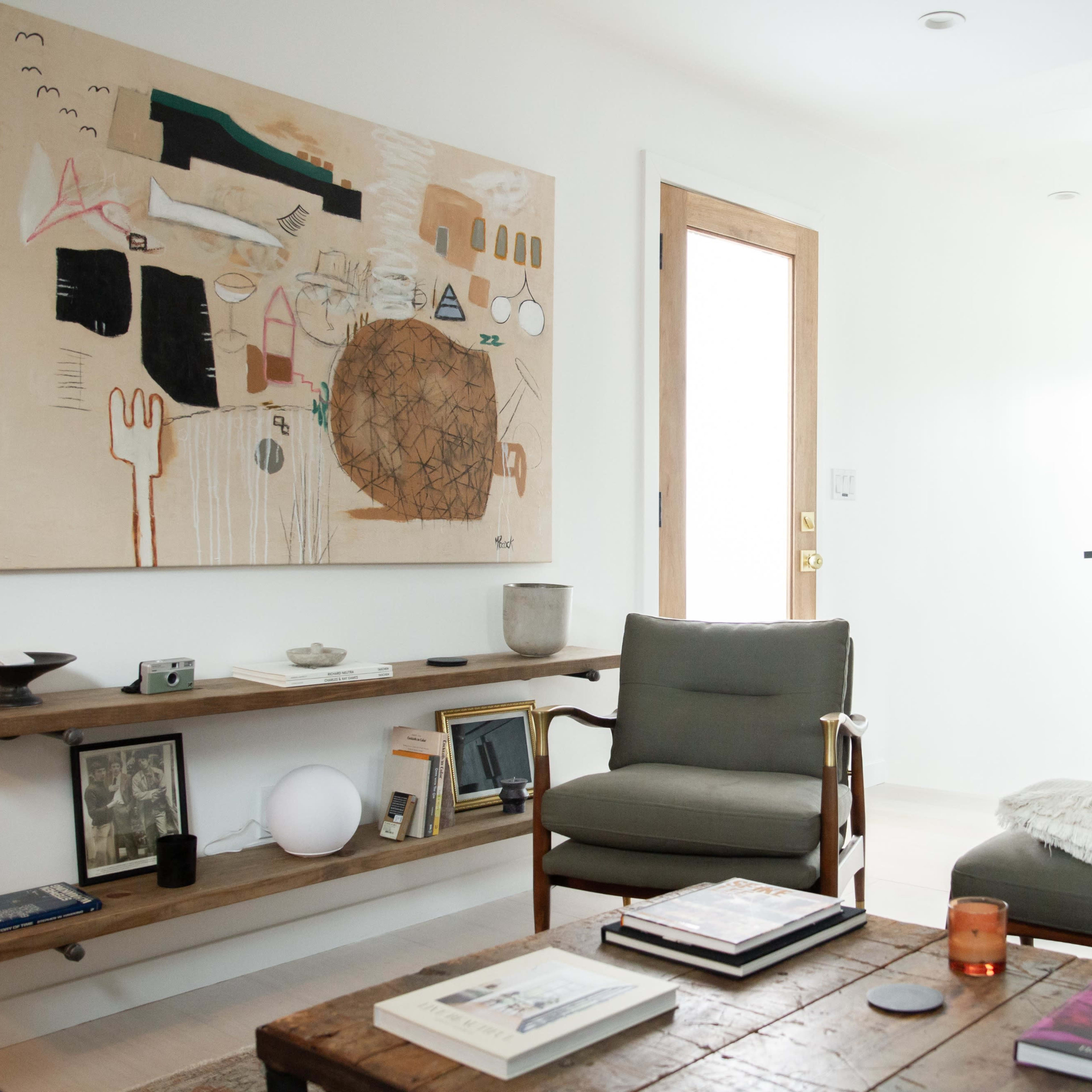 A modern living room features a textured painting on the wall, a wooden coffee table with books, two chairs, and a dark cabinet.