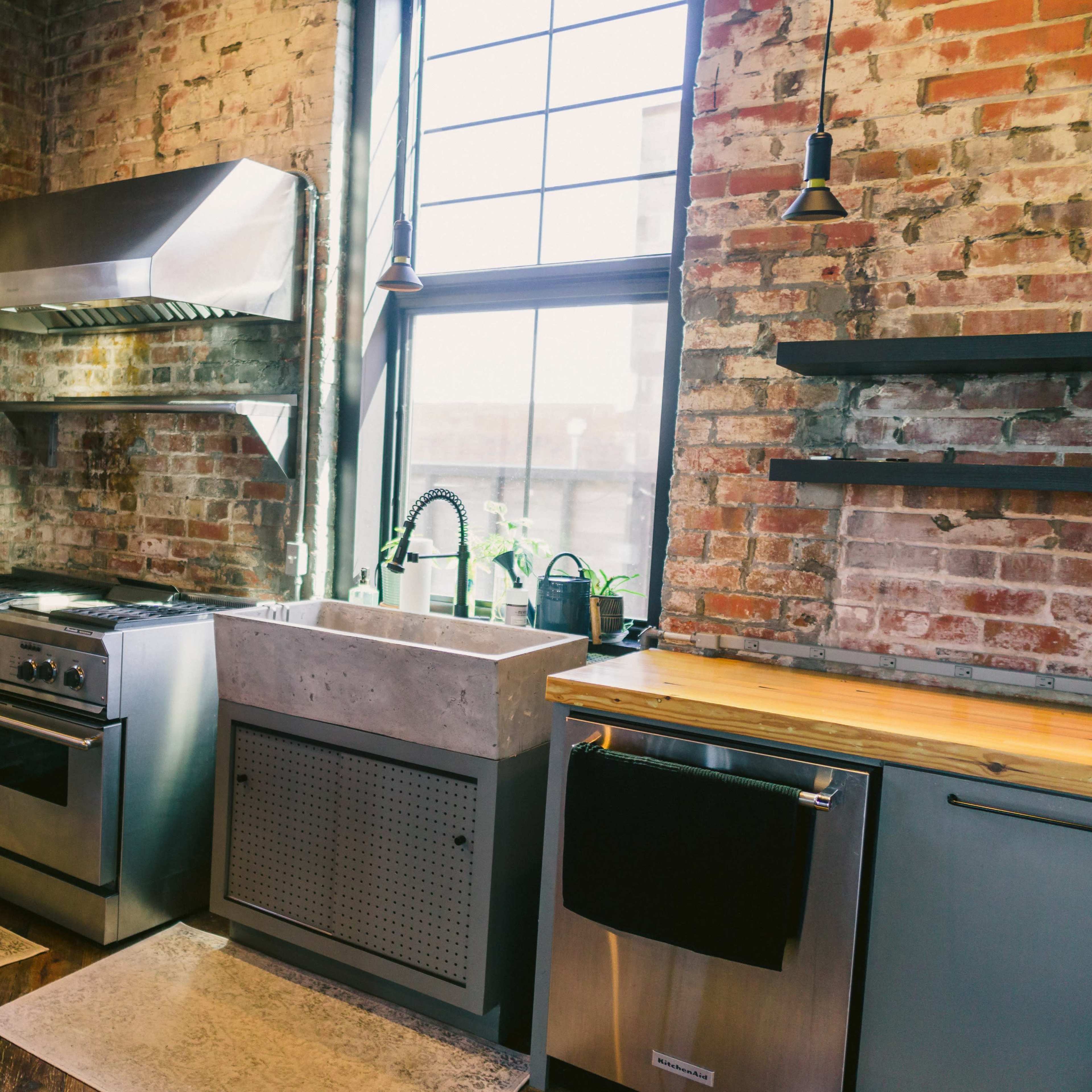 A kitchen with exposed brick walls, a stainless steel range hood above a gas stove, a concrete sink, and wooden countertops.