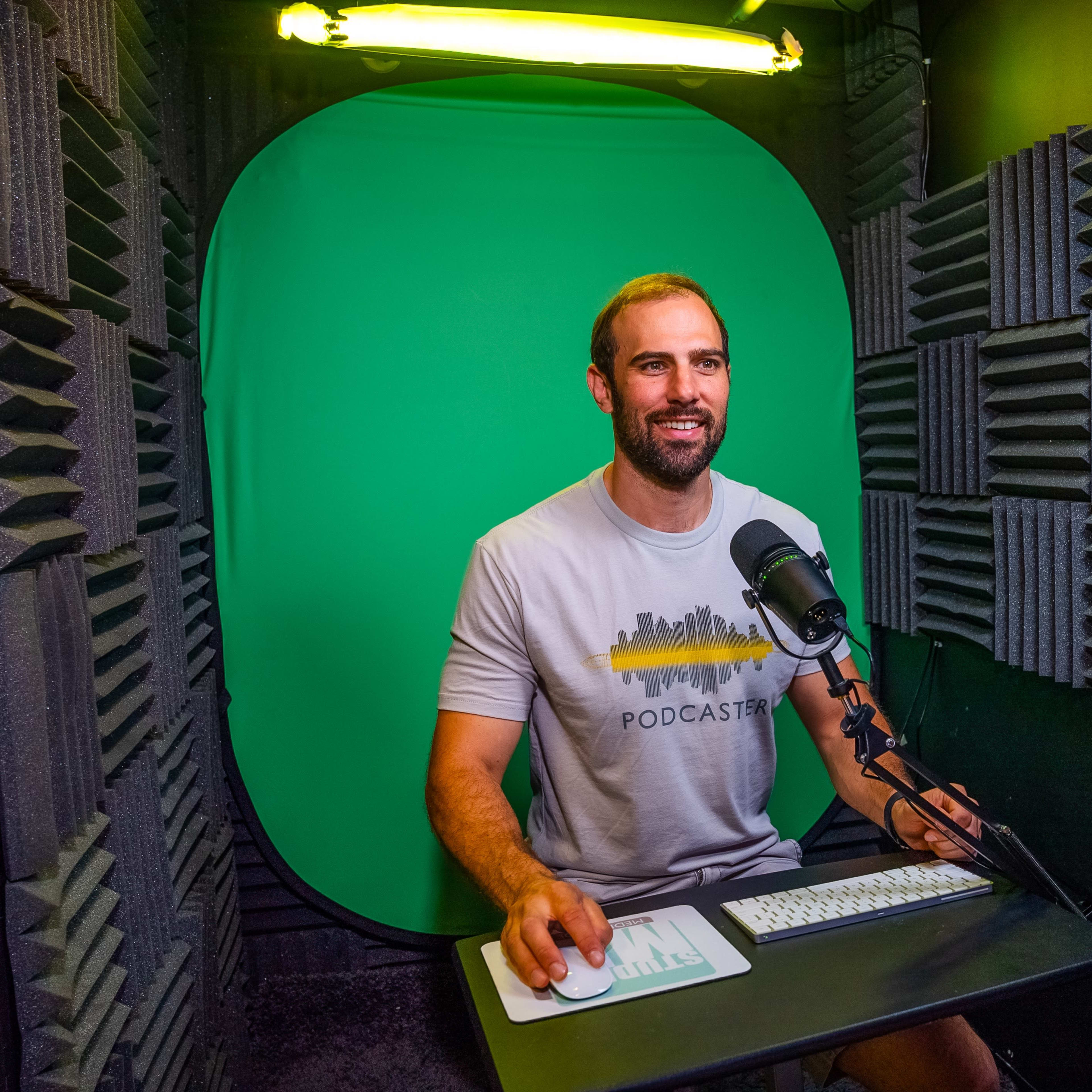 A man sits at a desk in a soundproof studio with a green backdrop, a microphone, and a computer keyboard.