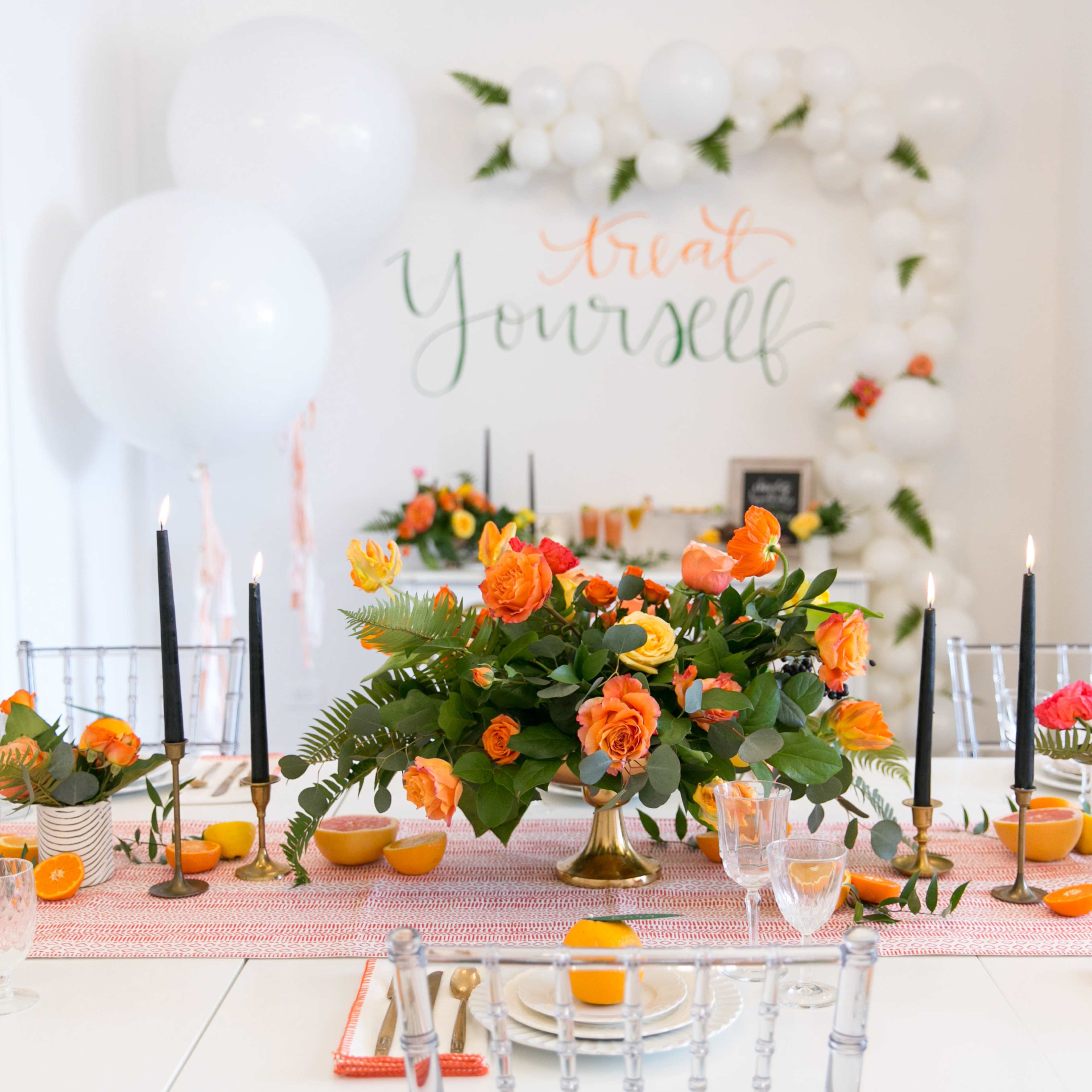 A decorated dining table features an orange floral centerpiece, black candlesticks, and vibrant place settings, set against a backdrop with the words "Treat Yourself."