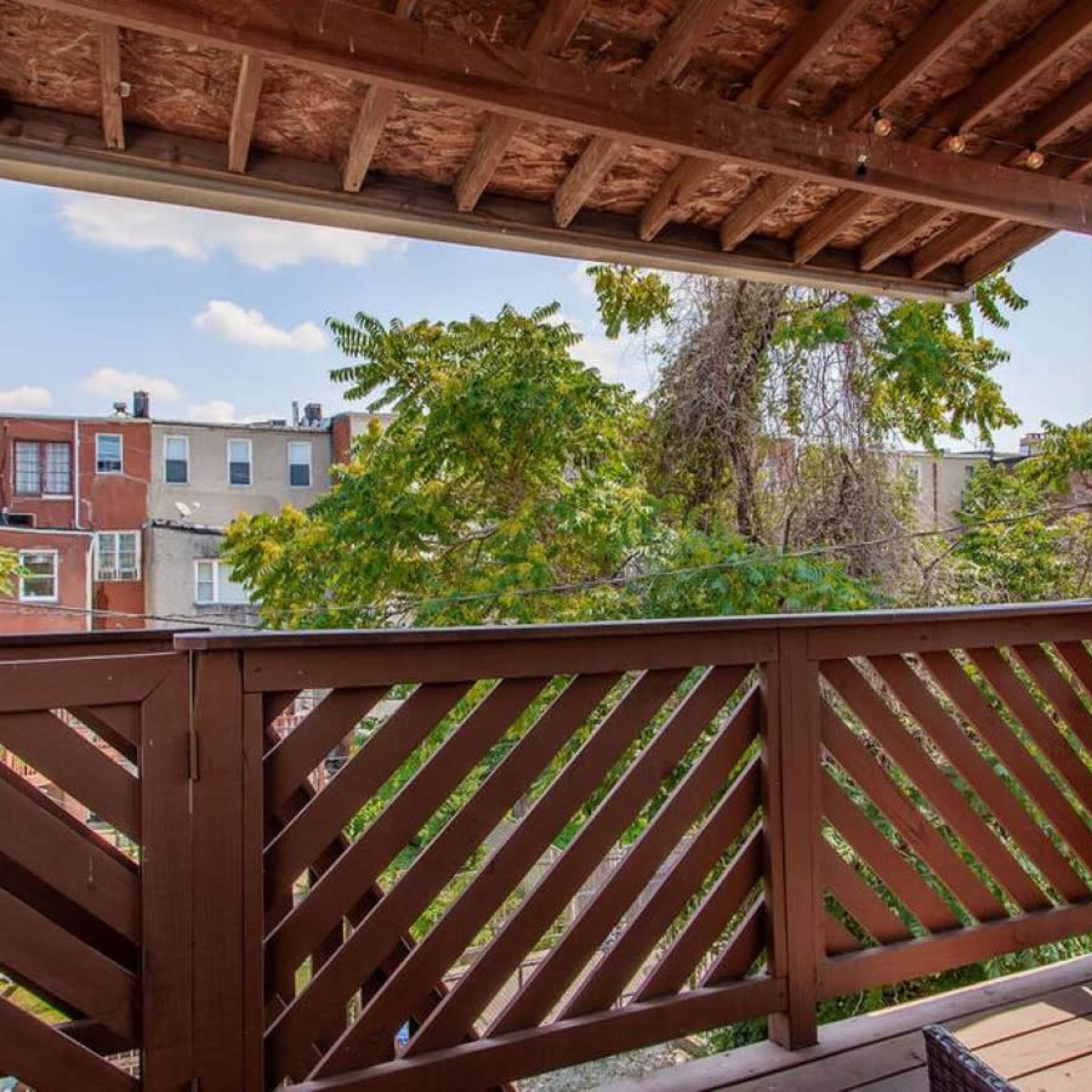A wooden balcony with a slatted railing overlooking green trees and buildings in the background.
