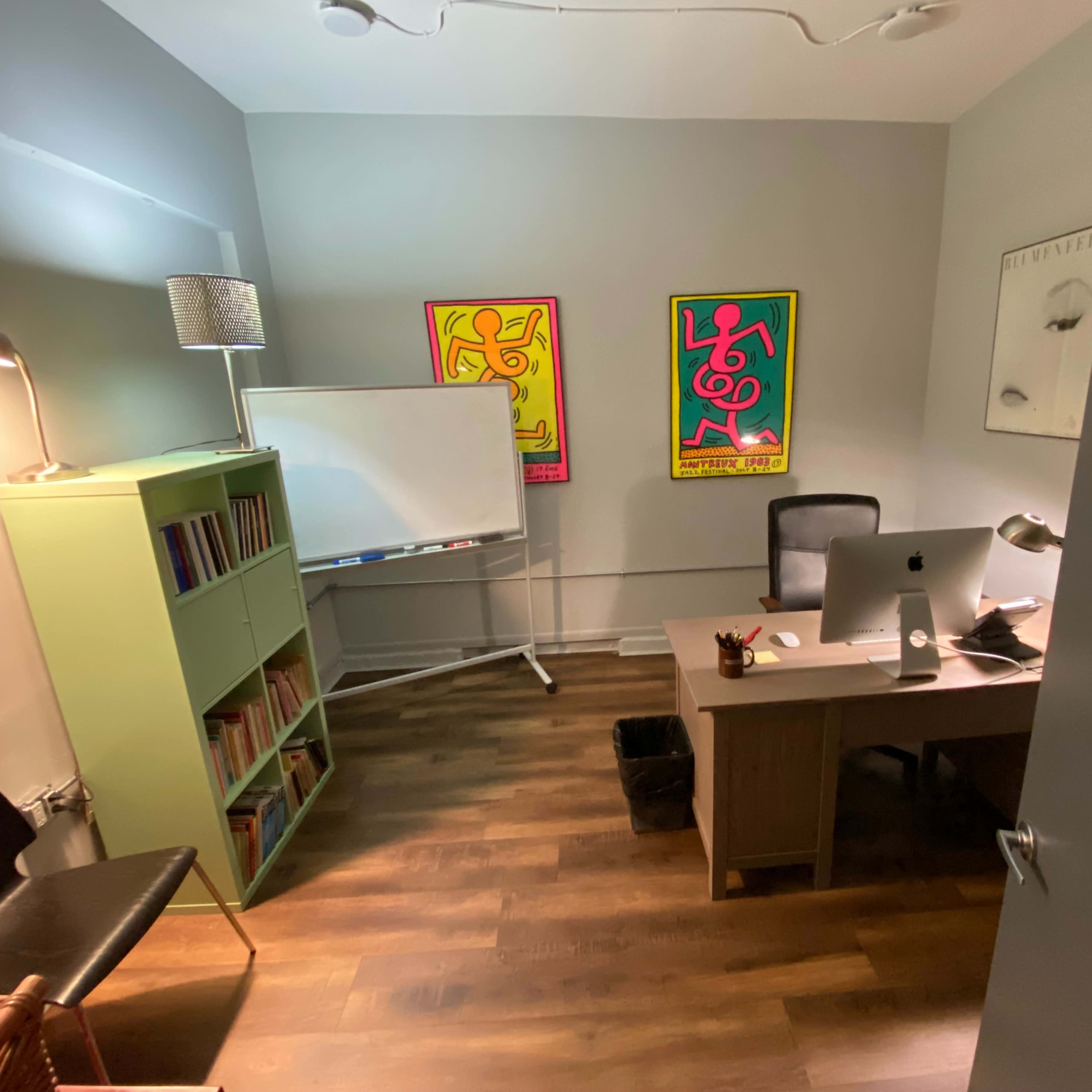 A small office features a desk, an ergonomic chair, a bookshelf filled with books, a whiteboard, and colorful artwork on the walls.