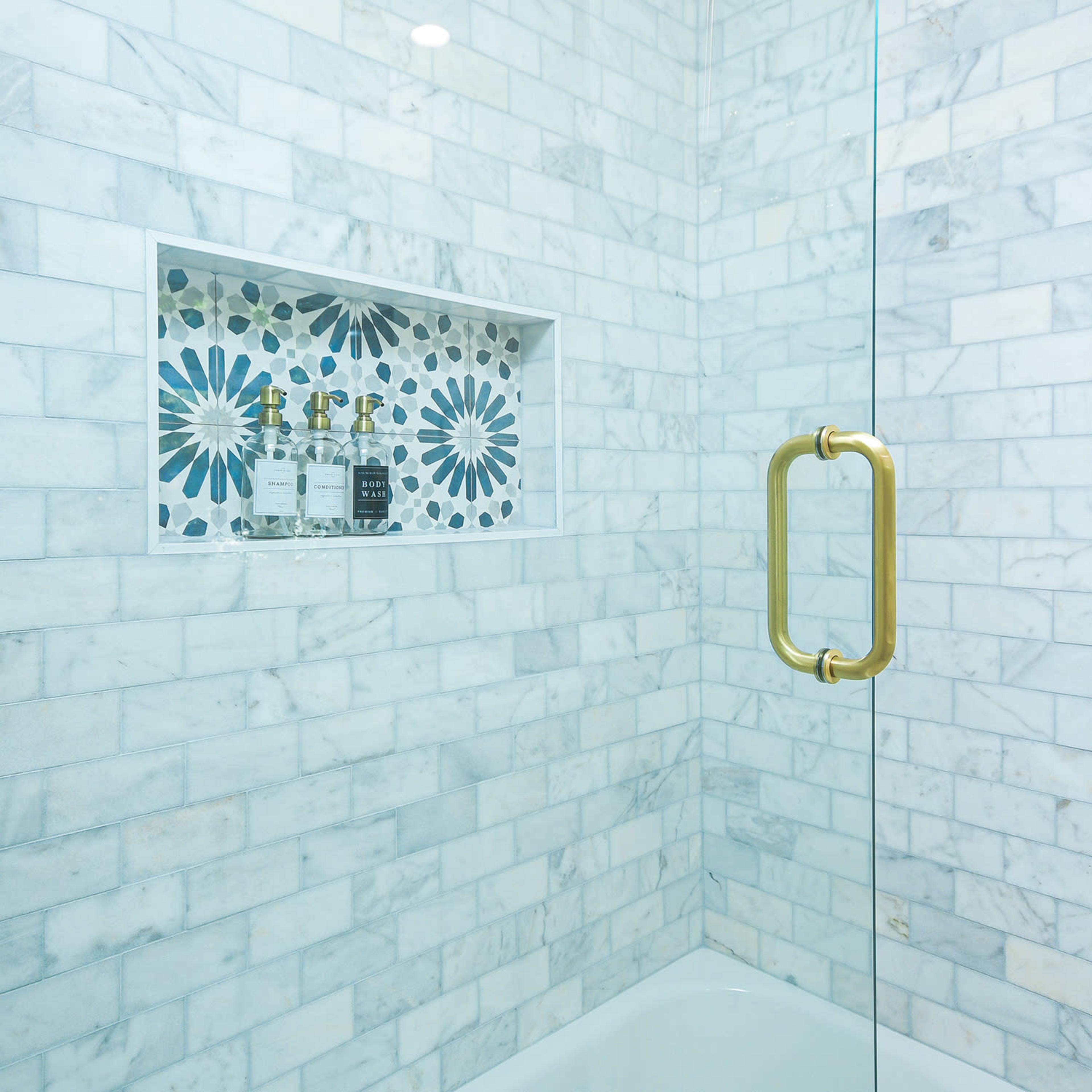 A modern shower with white marble tiles, a decorative niche featuring floral patterns, and a glass door with a gold handle.