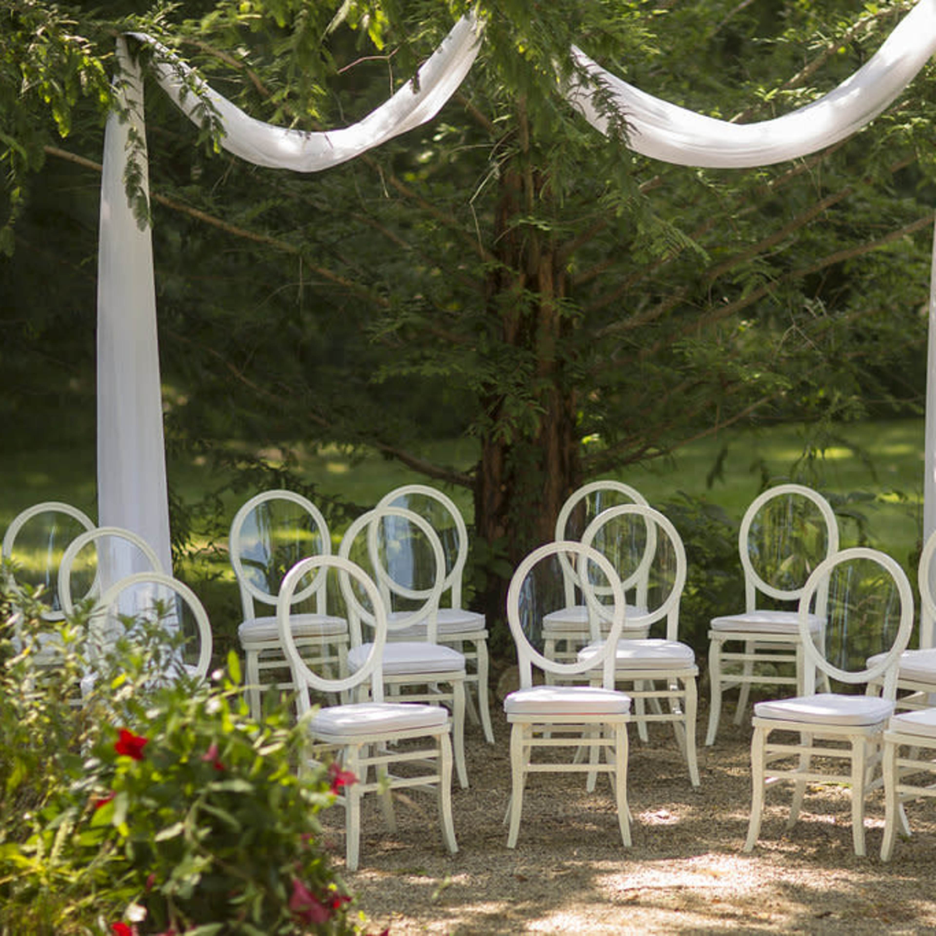 A cluster of white chairs is arranged beneath draped fabric and trees in a garden setting.