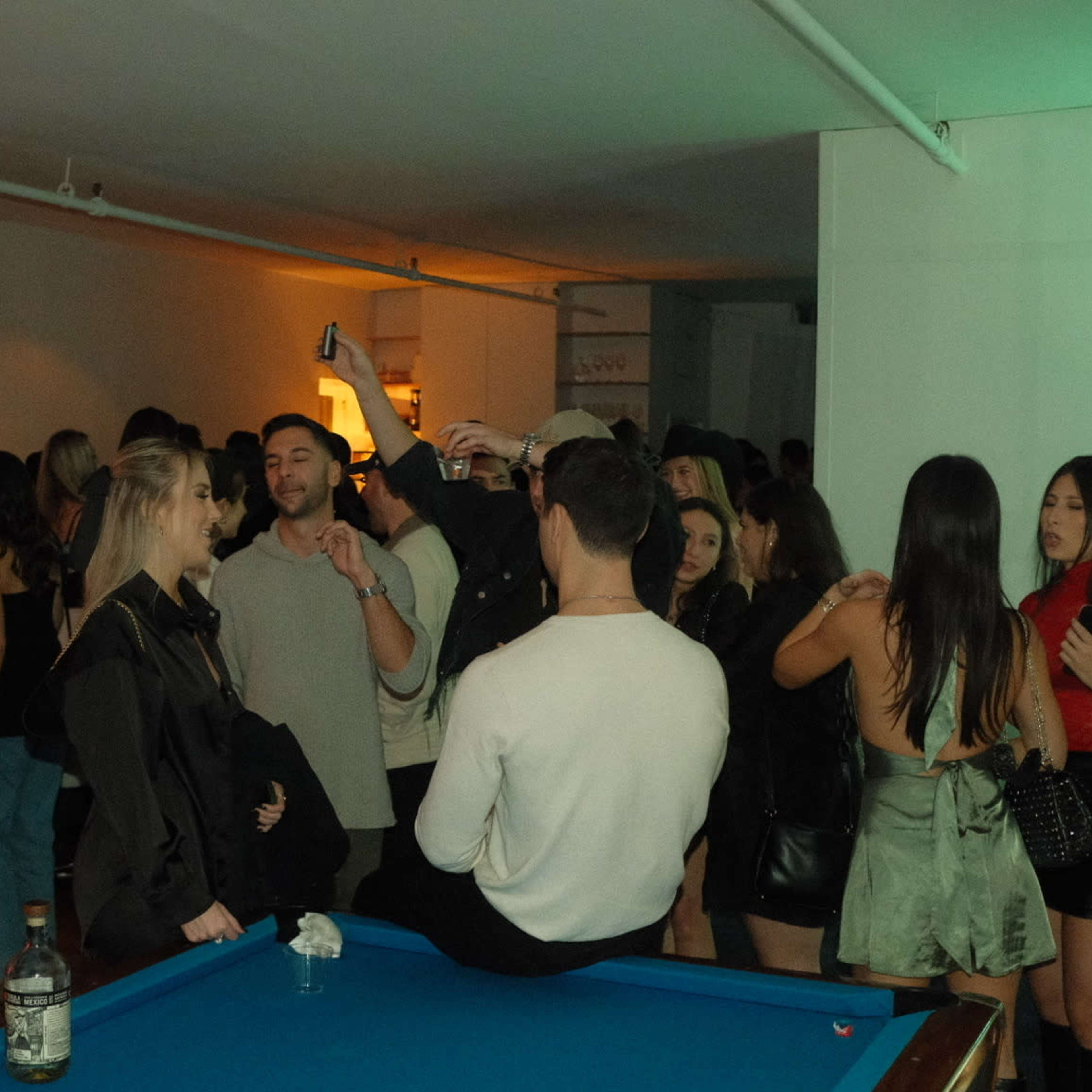 A crowded indoor party scene features groups of people socializing near a blue pool table, with a variety of drinks visible.