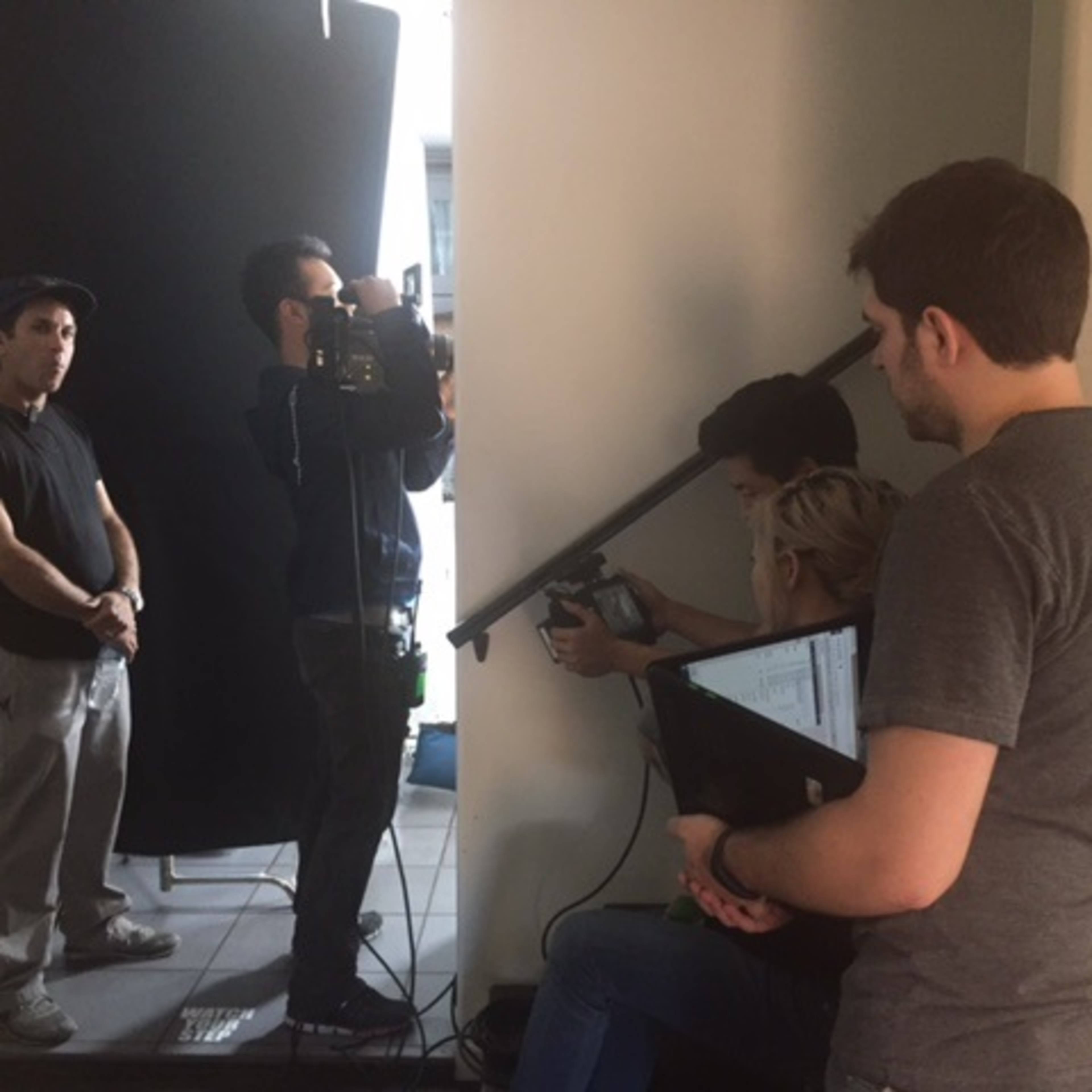 A small film crew is set up in a dimly lit indoor space, with one person operating a camera while others manage equipment and monitor the shot.
