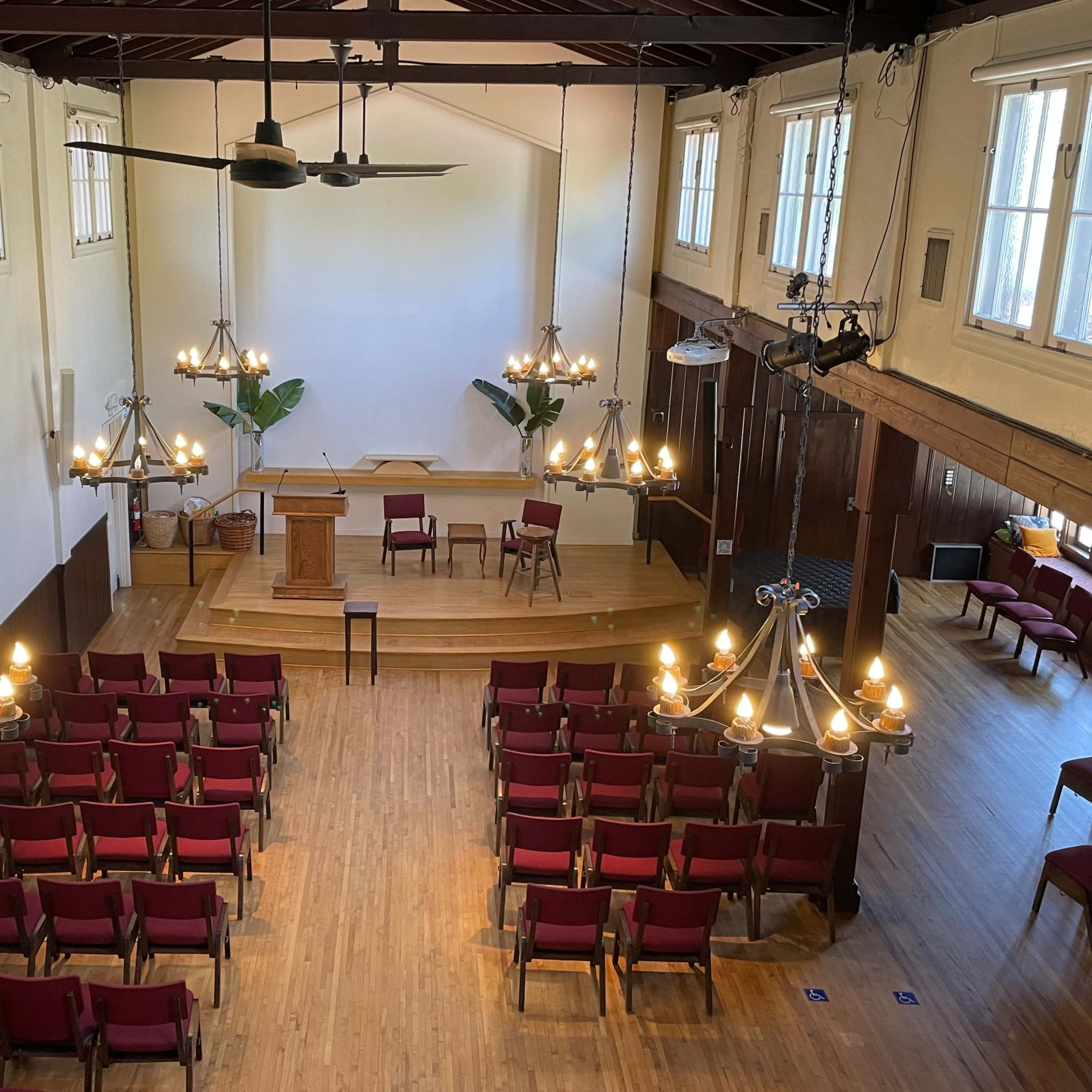 A spacious hall set up for a meeting, featuring rows of red chairs facing a wooden podium and two chairs on a raised platform, with chandeliers hanging from the ceiling.