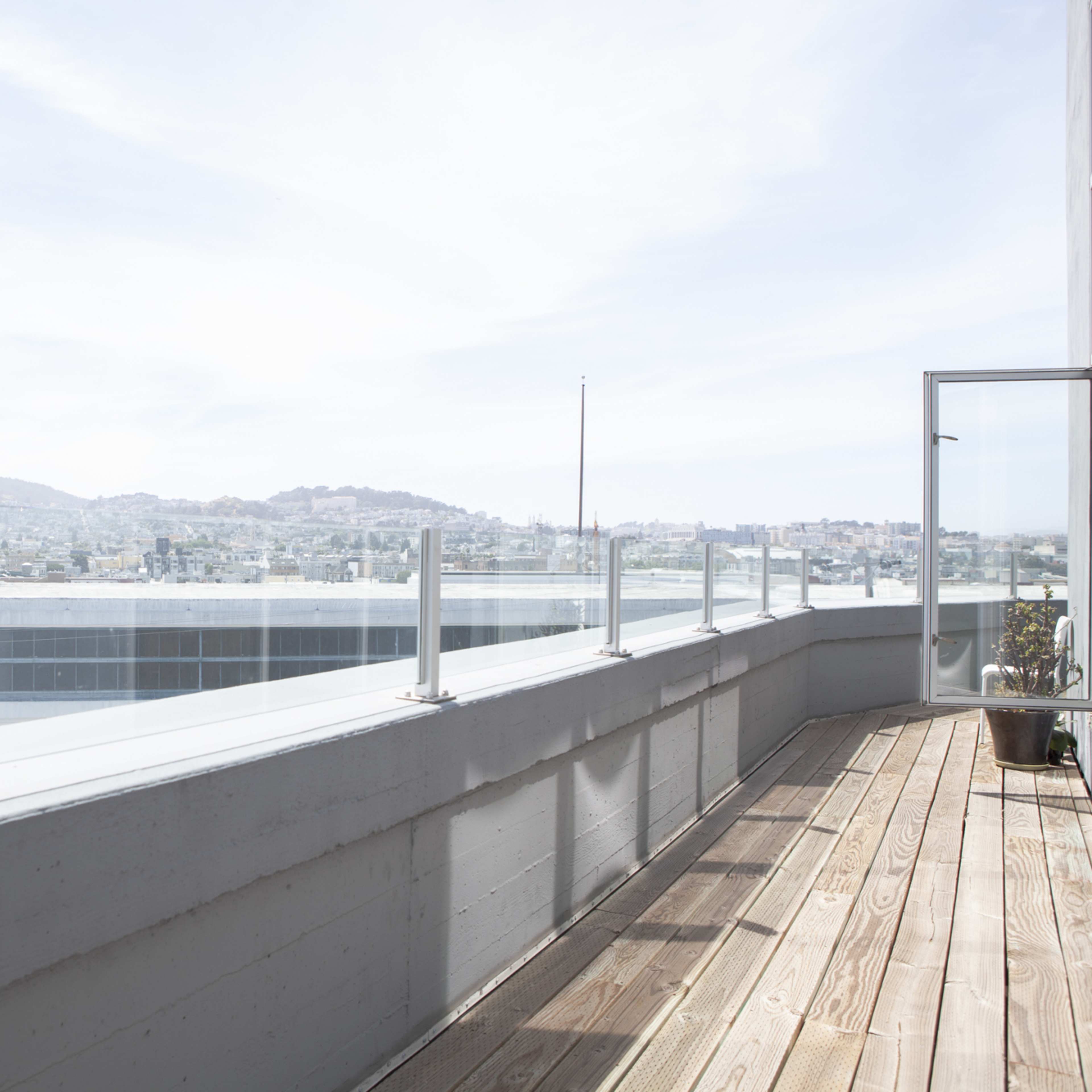 A spacious balcony with wooden flooring overlooks a cityscape and distant hills under a clear sky.