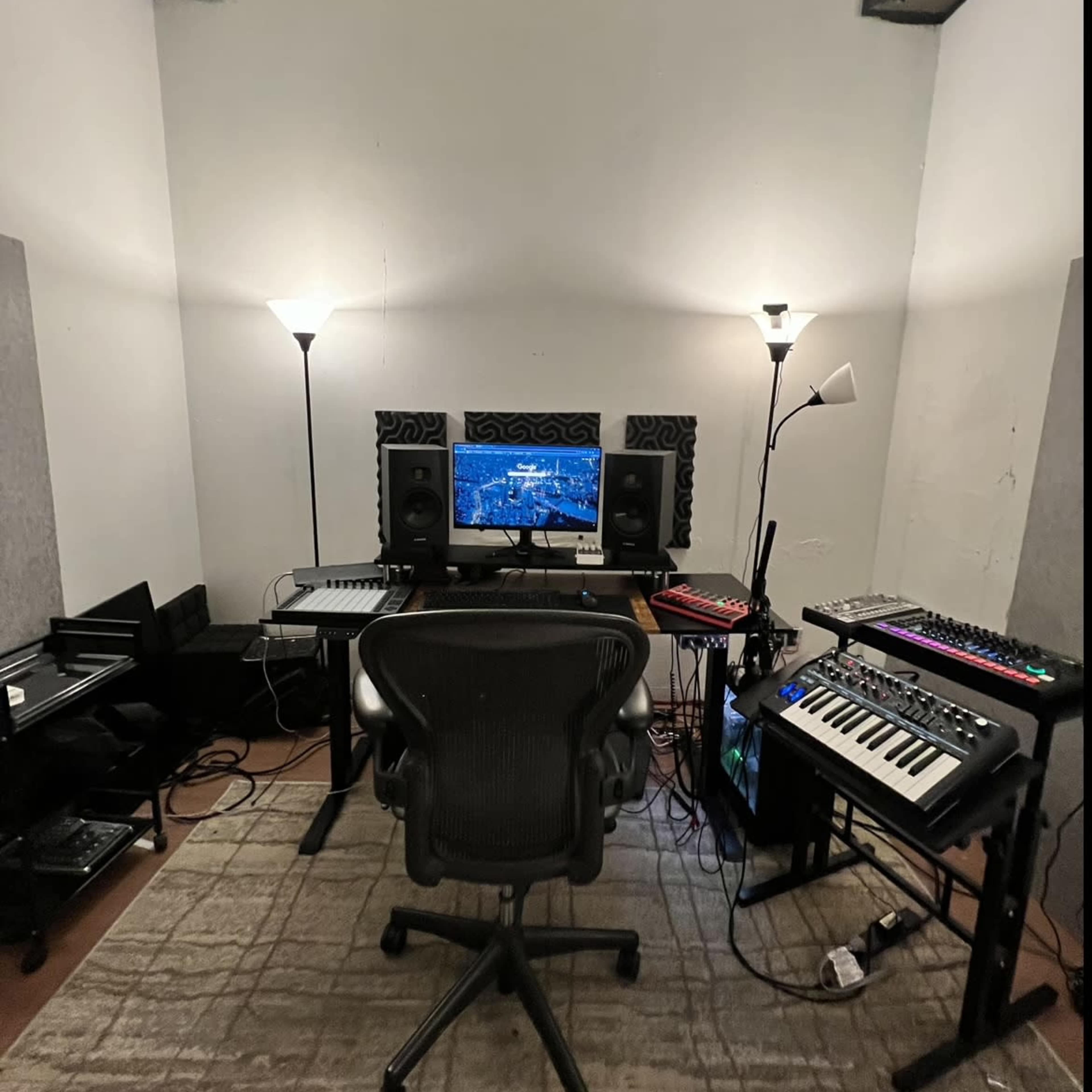 A music production studio is shown, featuring a desk with a computer monitor, speakers, a keyboard, and various electronic equipment, all positioned around a central office chair.