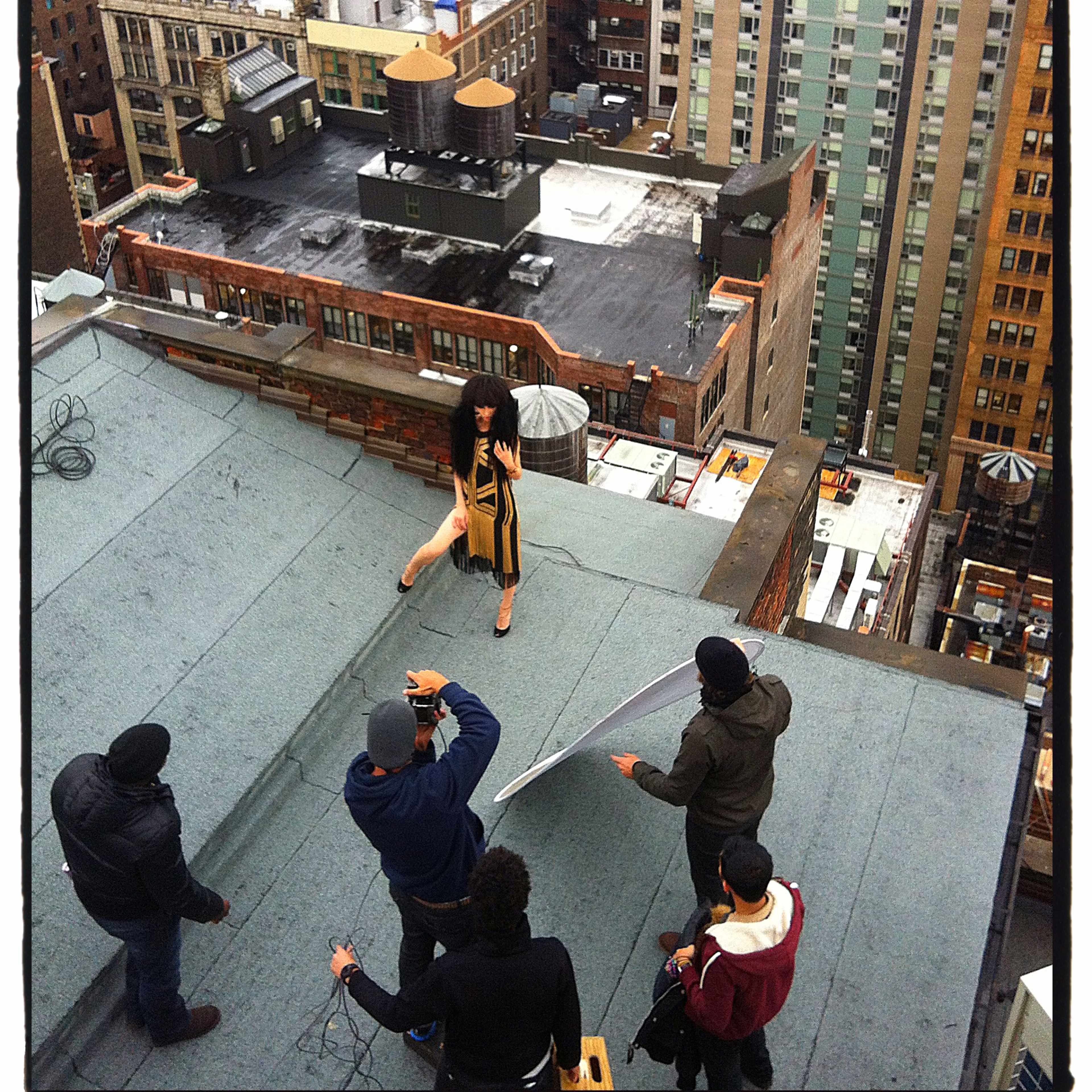 A model poses for a photoshoot on a rooftop in an urban setting while a small crew adjusts lighting and equipment.