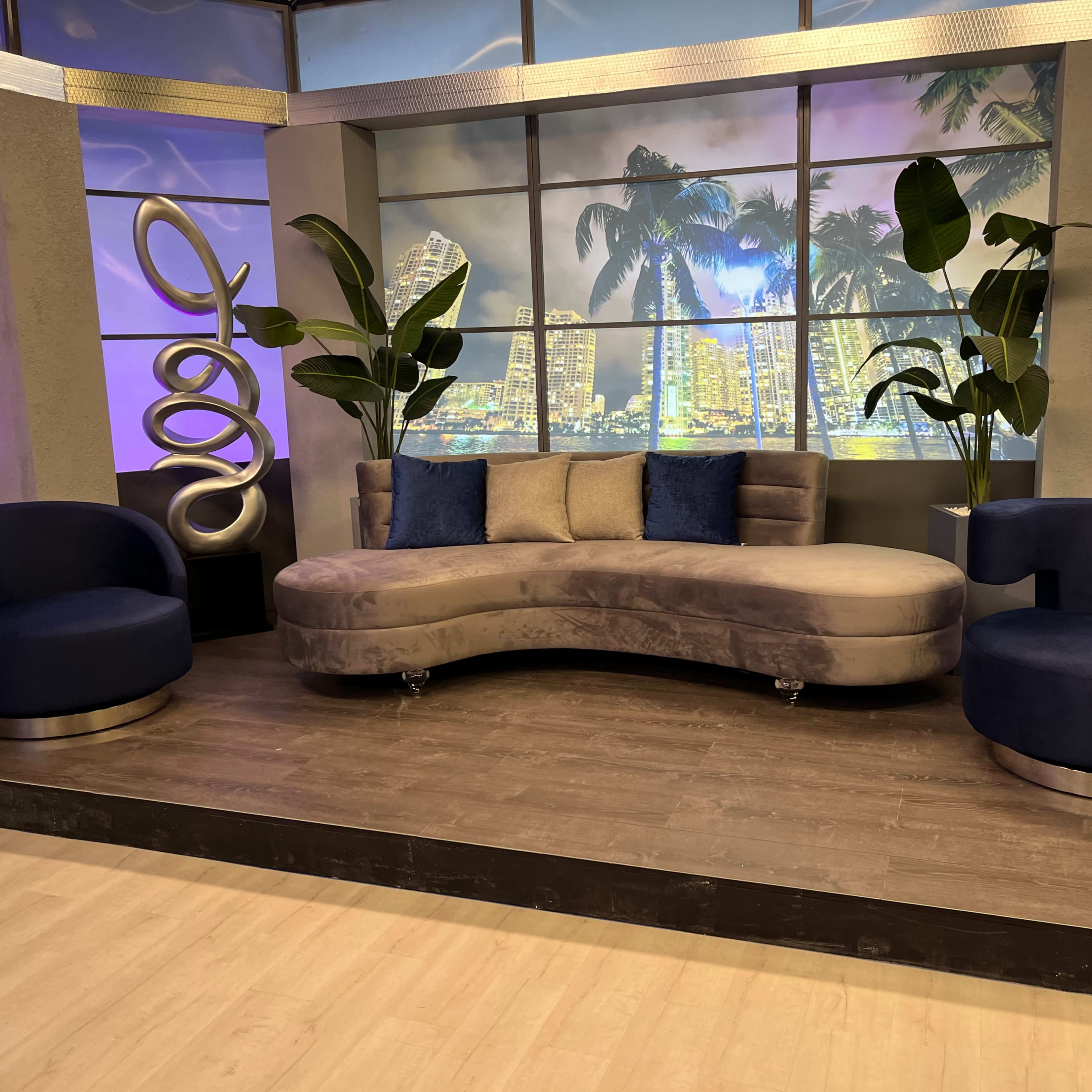 A modern television set featuring a curved gray sofa flanked by two dark blue chairs, with large potted plants and a city skyline backdrop.