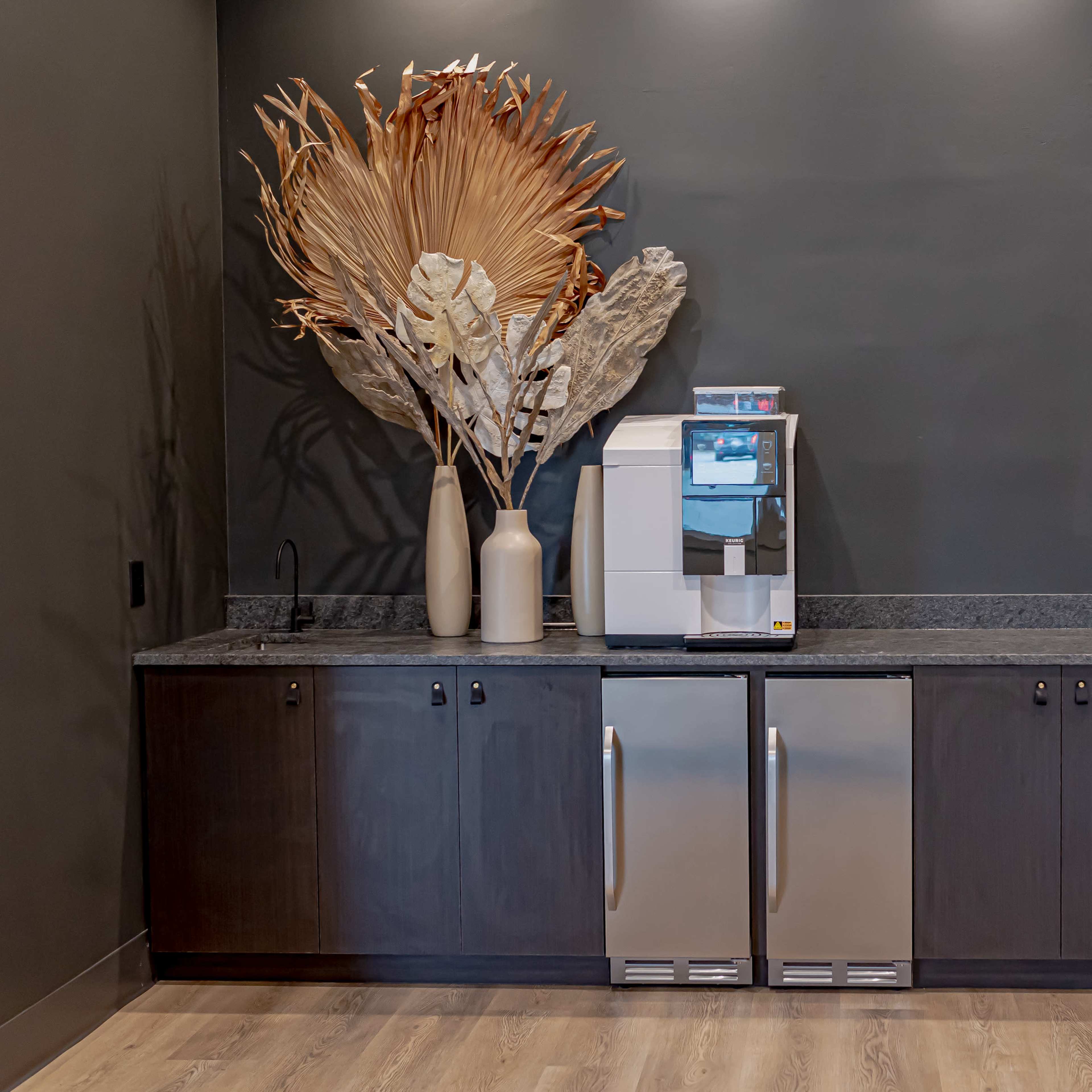 A modern kitchenette featuring a drink dispenser, two stainless steel refrigerators, and large decorative dried plants on a countertop.