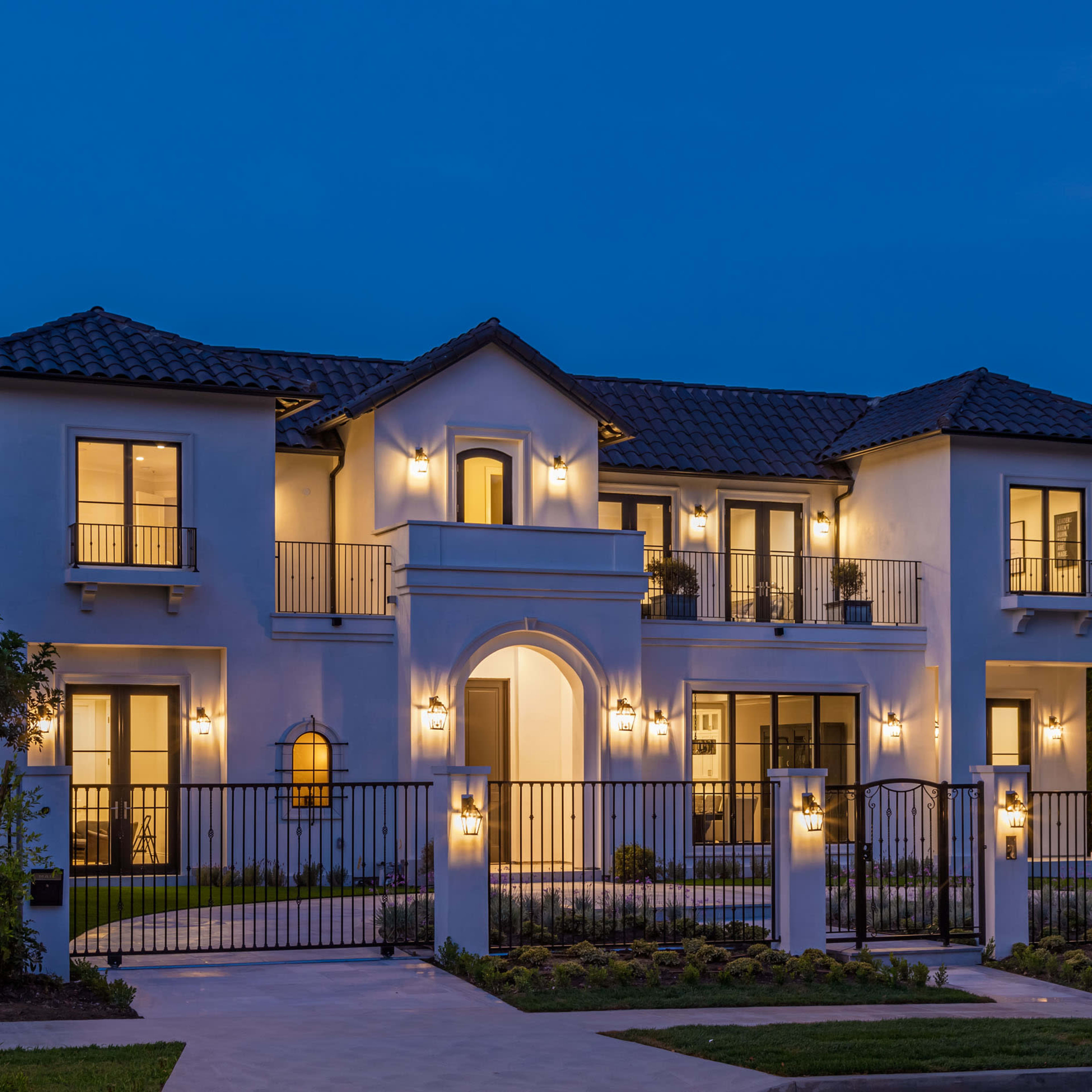 A two-story white stucco house with a tiled roof is illuminated by outdoor lights and surrounded by a wrought iron fence.