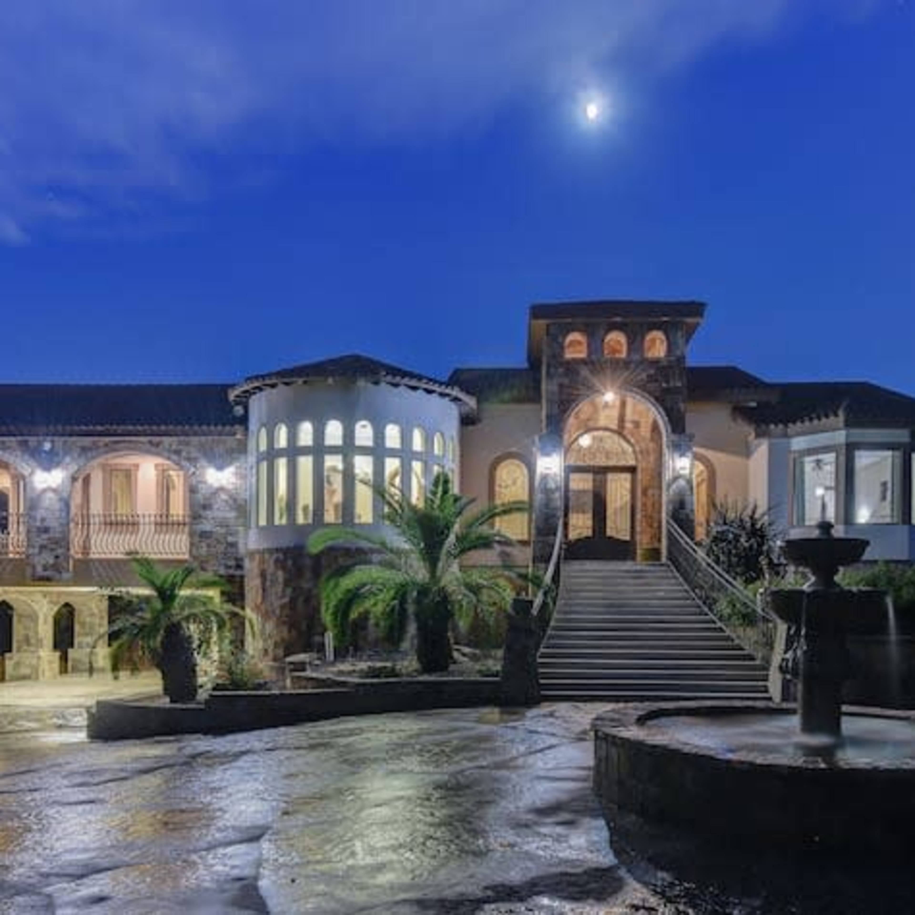 A large, stone-fronted mansion with multiple balconies and a fountain in the foreground is illuminated under a night sky.