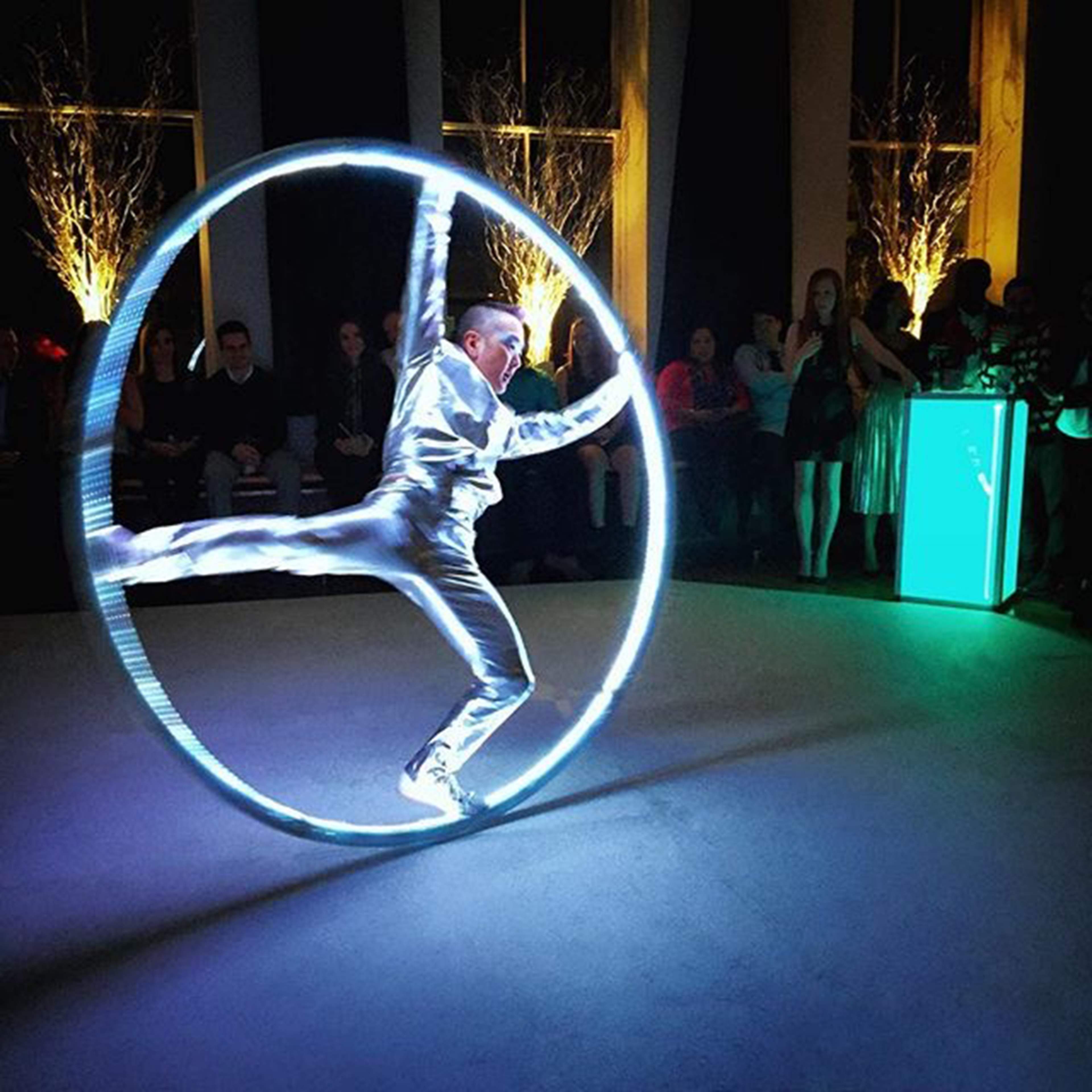 A performer dressed in silver is executing a maneuver inside a glowing circular apparatus at a venue filled with people.