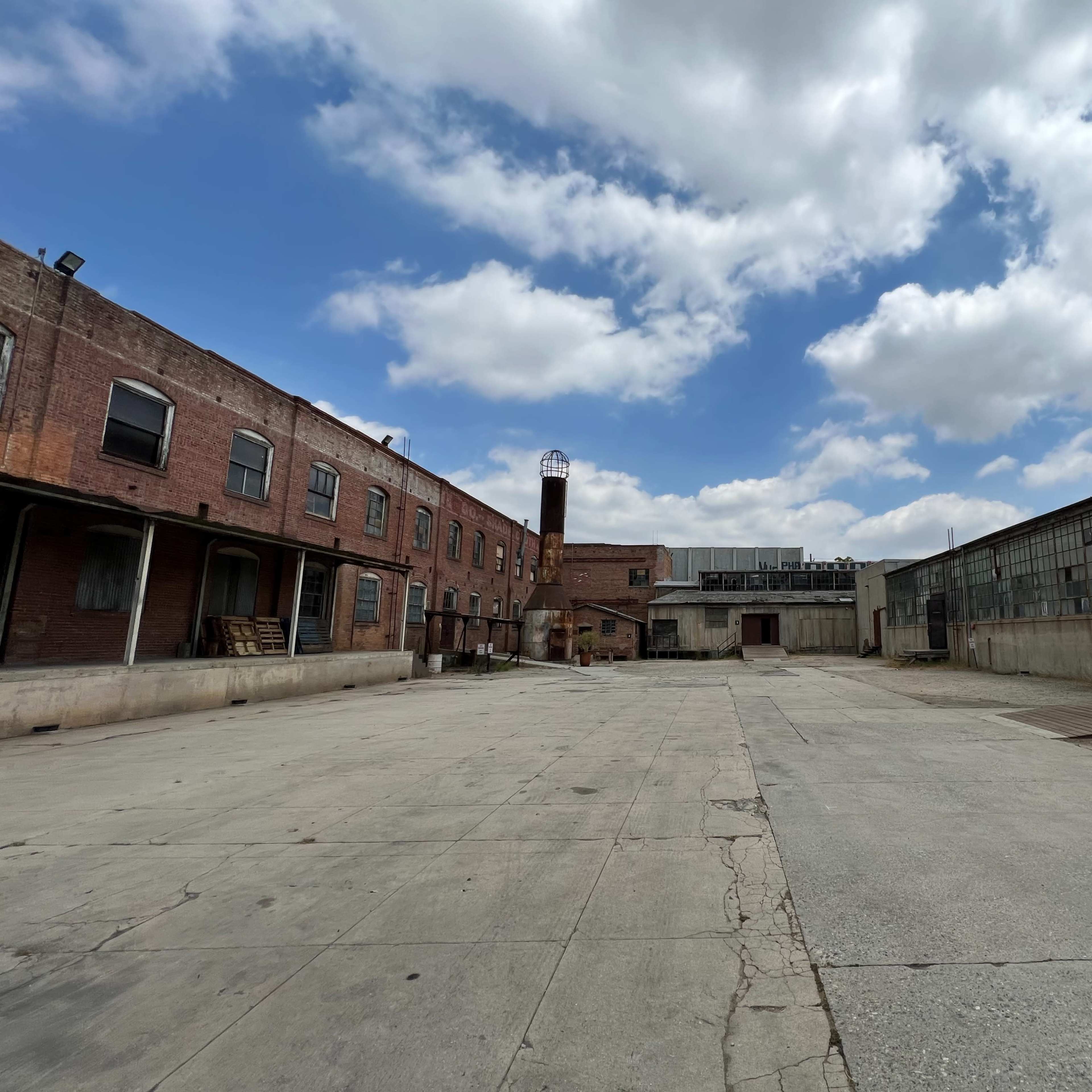An empty industrial yard surrounded by brick buildings and large metal structures under a cloudy sky.