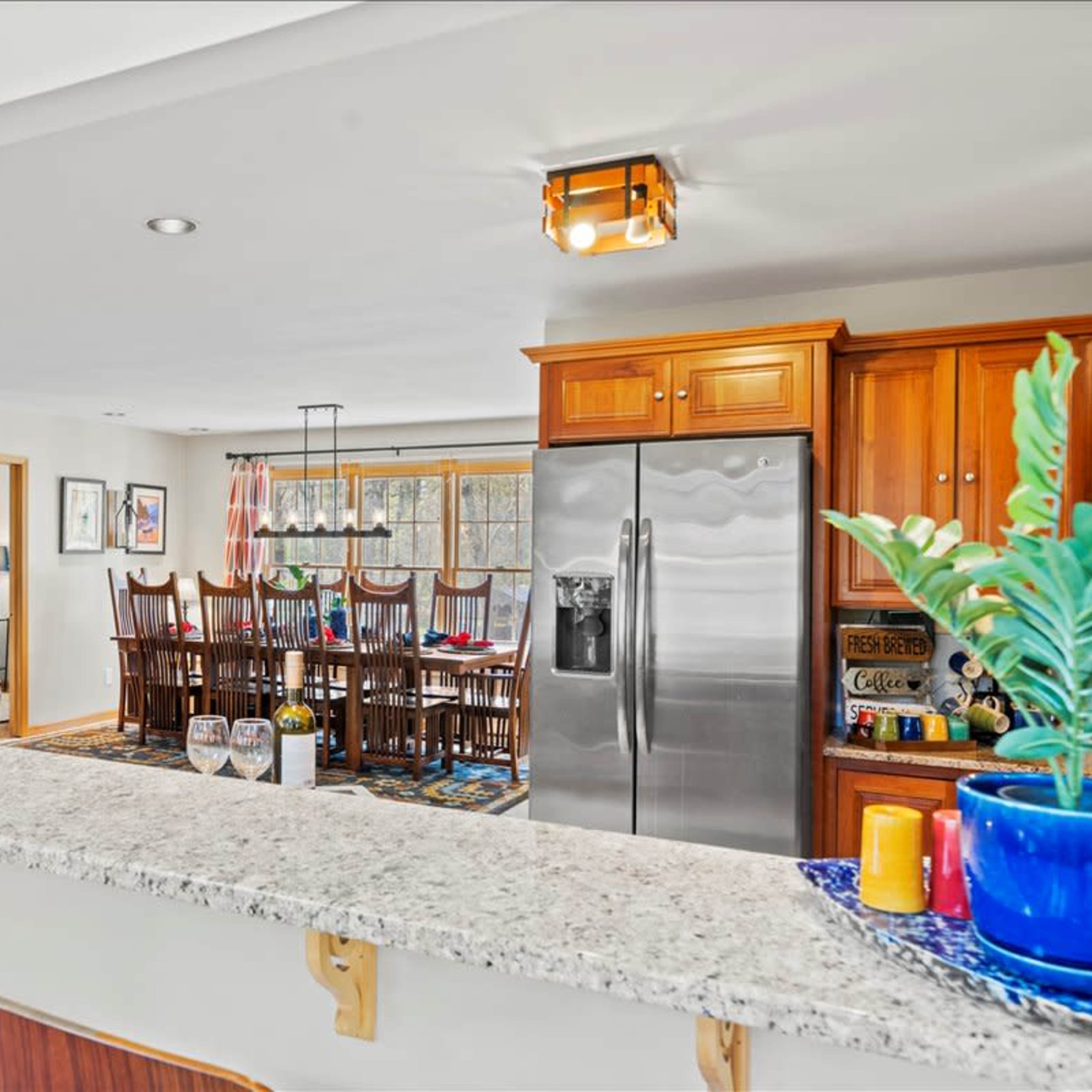 A kitchen with wooden cabinets and a granite countertop opens to a dining area featuring a large table surrounded by chairs.