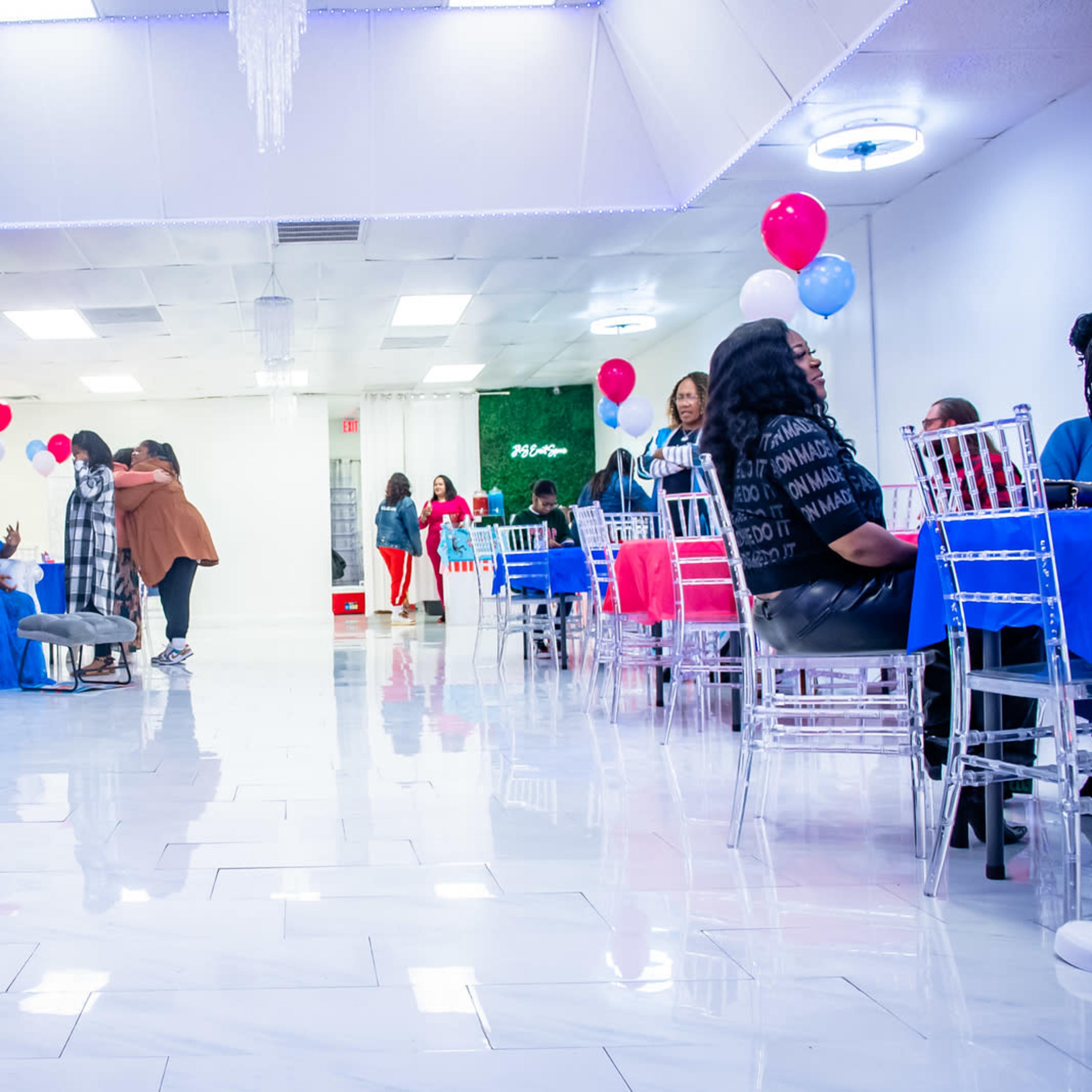 A brightly lit event space decorated with blue and red balloons, featuring tables with chairs set up for guests, some of whom are mingling and socializing.