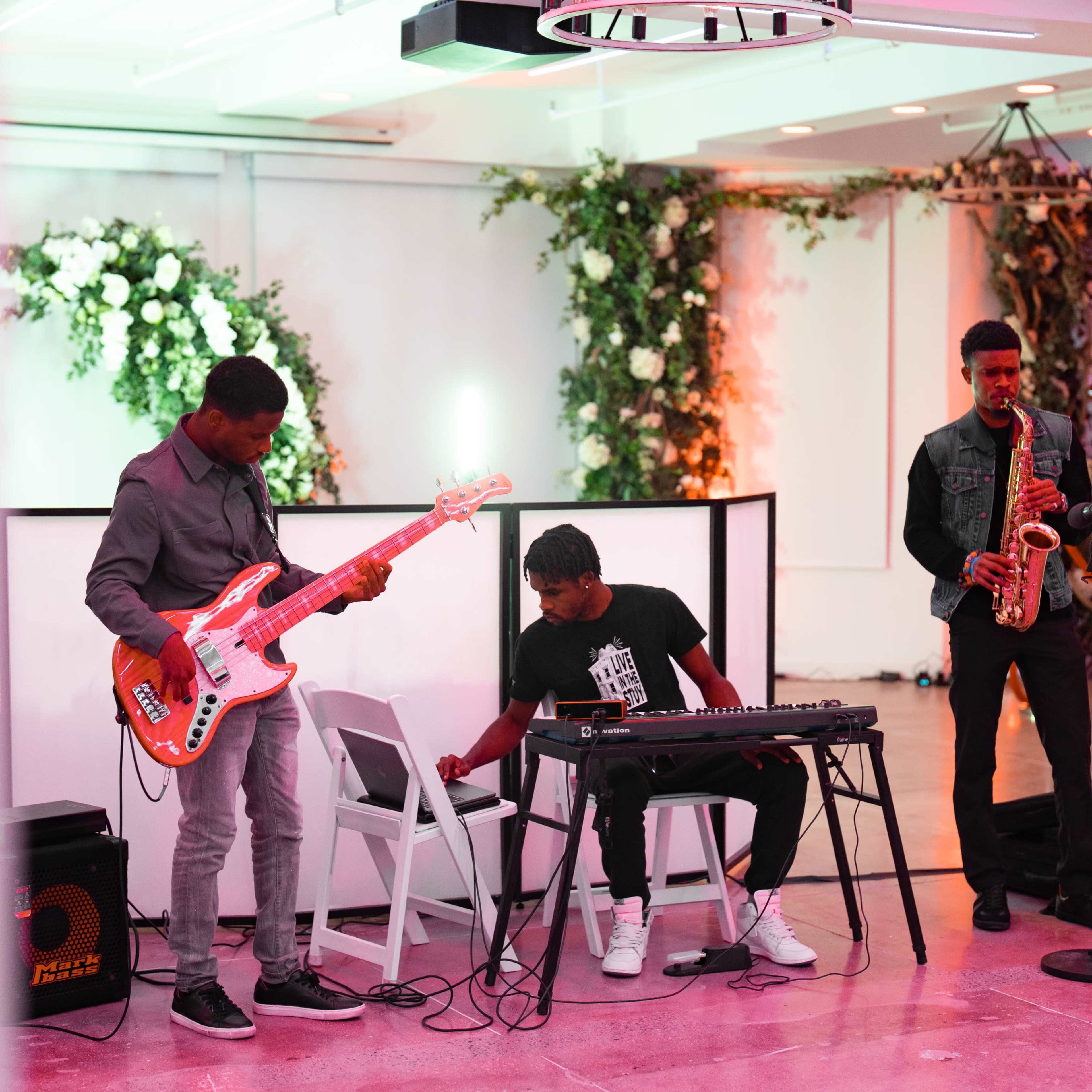 Three musicians perform in a brightly lit venue, with one playing a saxophone, another on a keyboard, and a third on an electric bass guitar.
