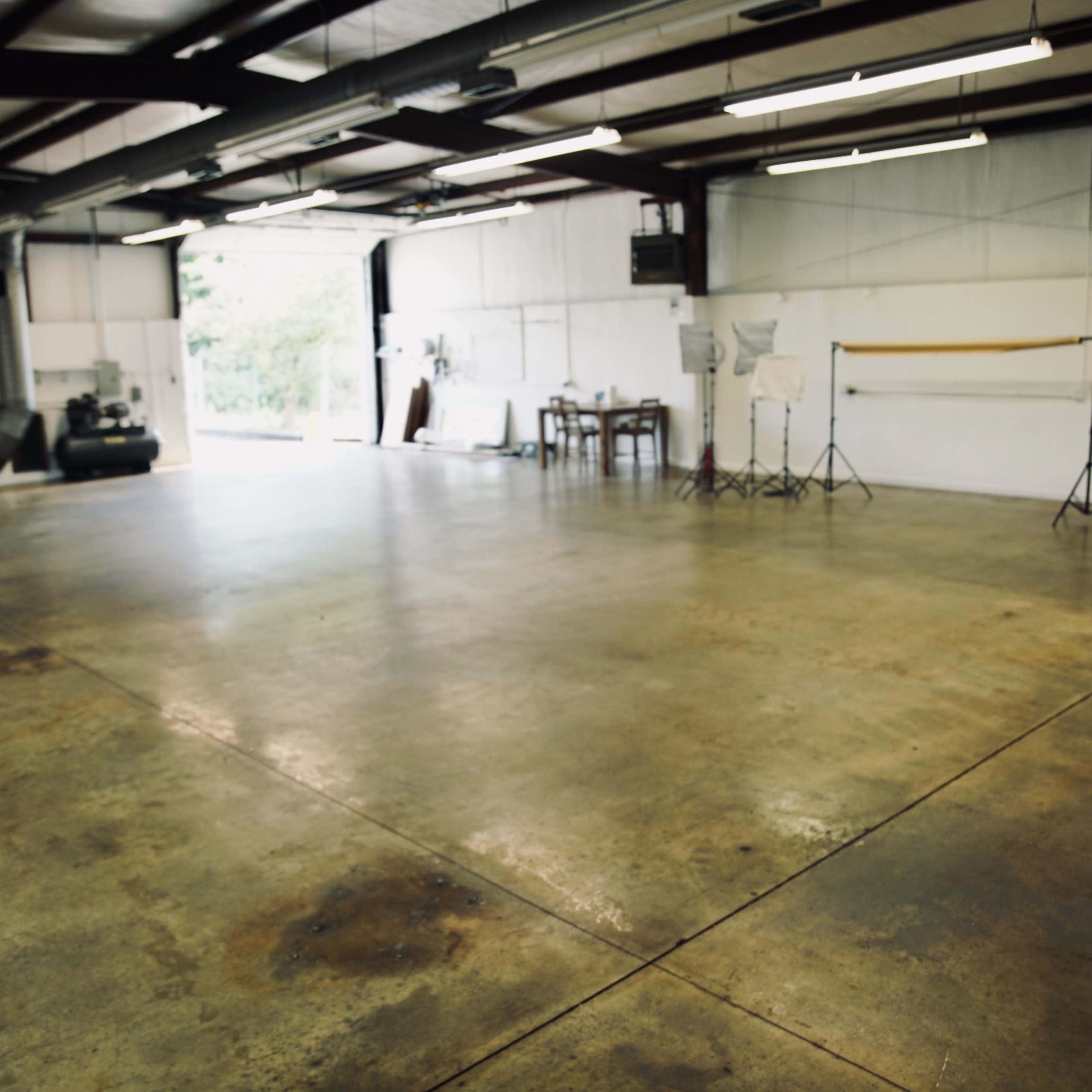 A spacious, empty warehouse with concrete floors and sparse equipment, including a table and photography stands, set against a backdrop of white walls.