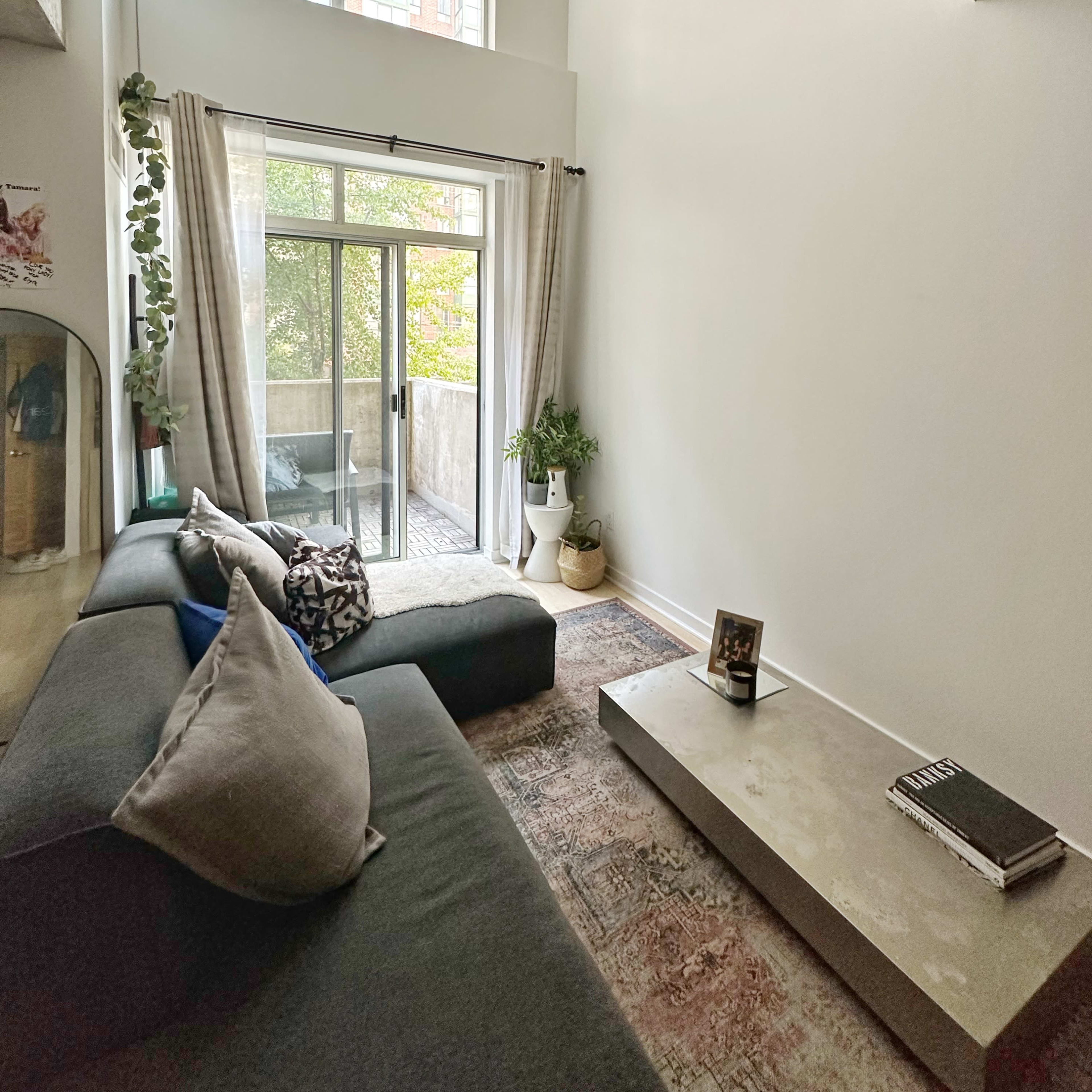 A modern living room with a black sofa, a concrete coffee table, large windows, and a balcony overlooking greenery.