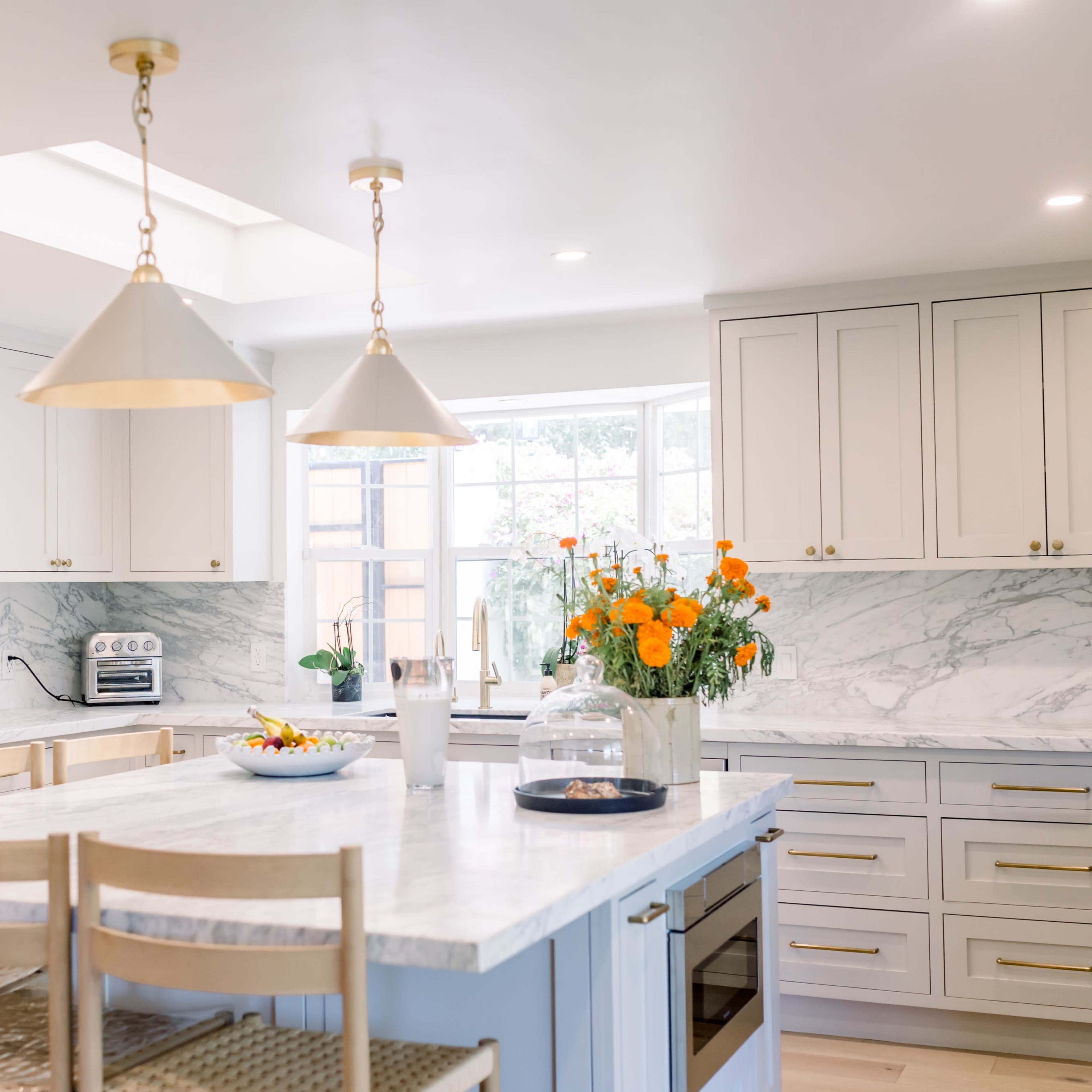 A modern kitchen featuring white cabinets, a marble countertop island, and a large window with a bouquet of orange flowers on the counter.