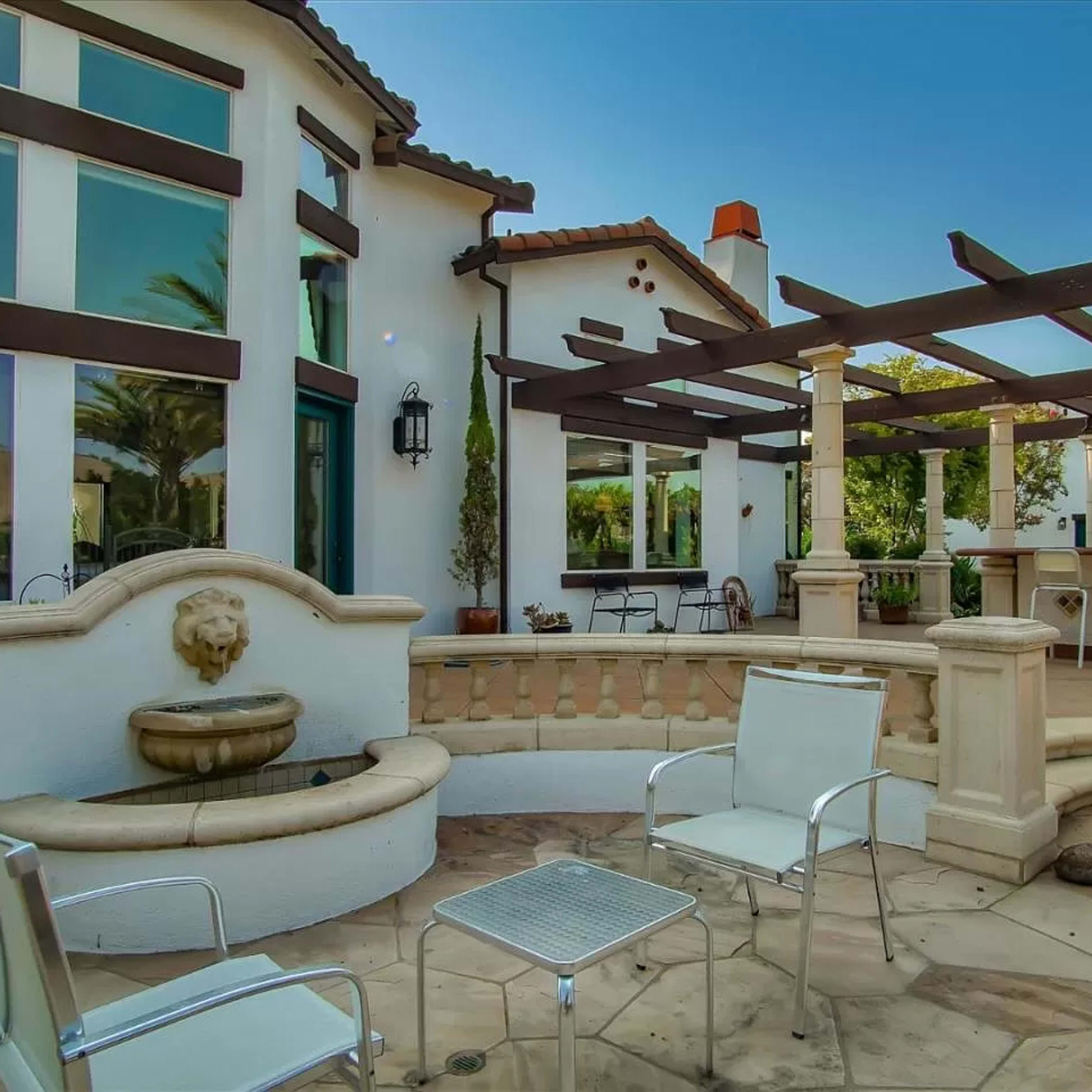 A landscaped outdoor patio area with a fountain, seating chairs, and a pergola near a residential building.
