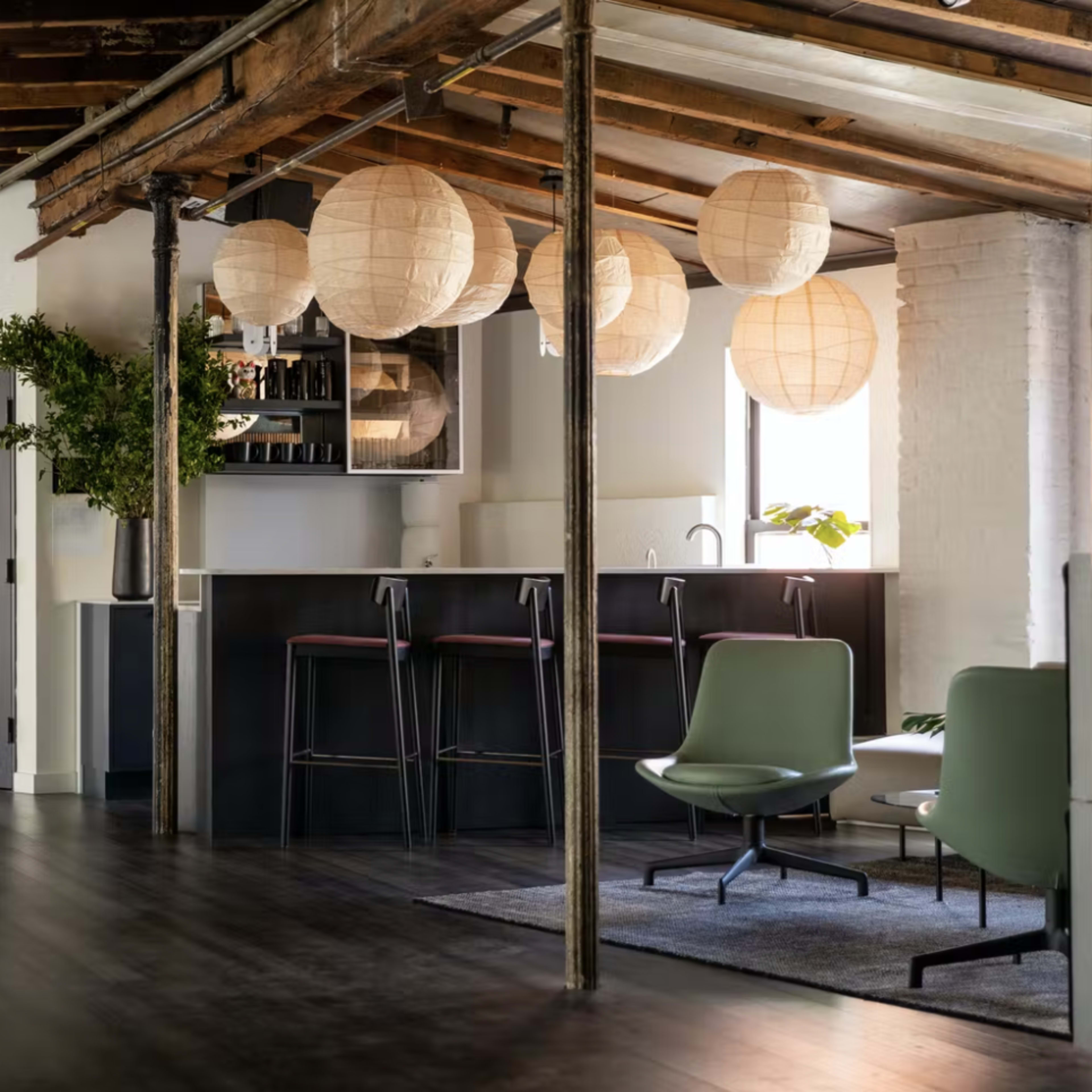 A modern, open-plan office space featuring a bar area with hanging lantern lights, green seating, and large windows.