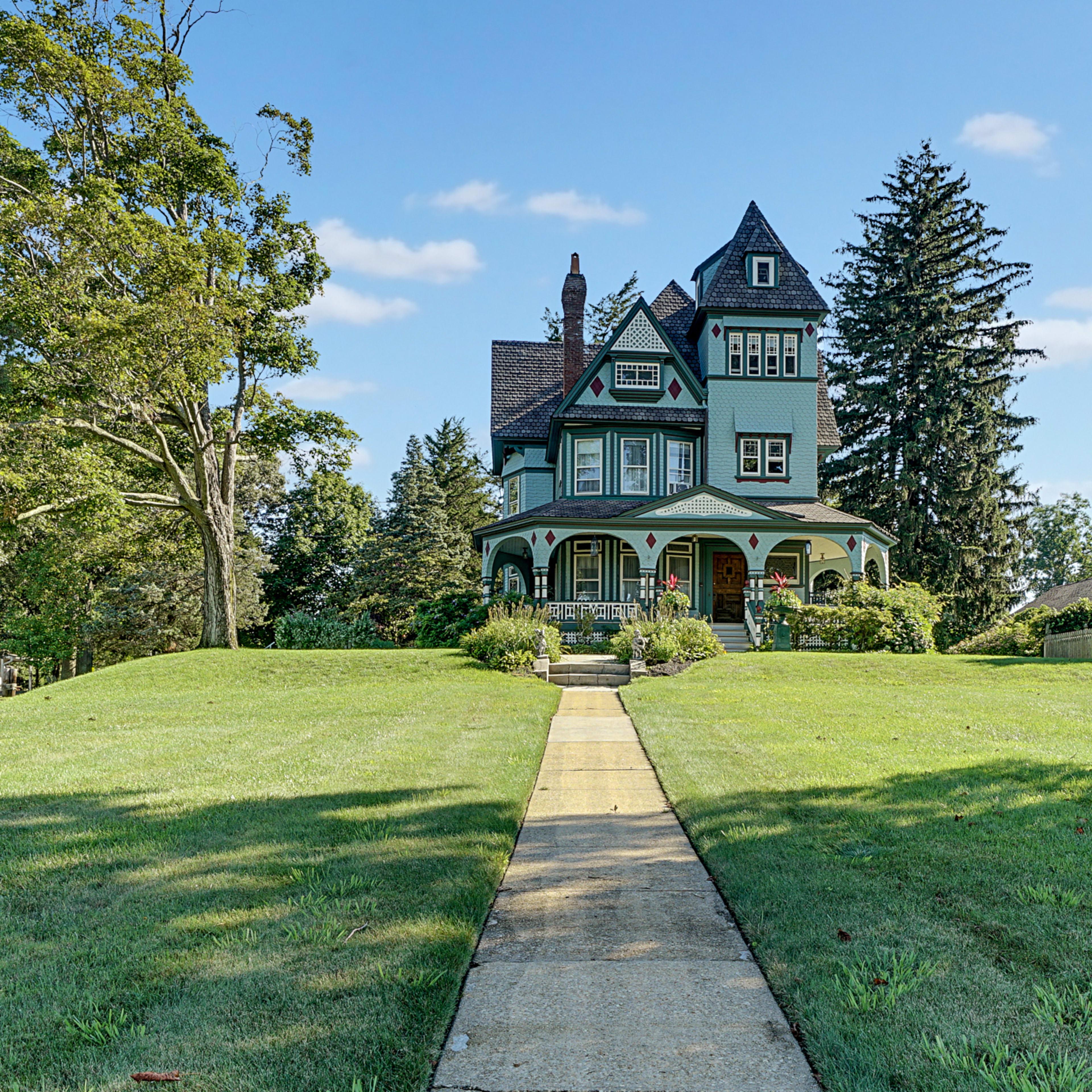 A large, green Victorian-style house stands at the end of a concrete path, surrounded by trees and a manicured lawn.