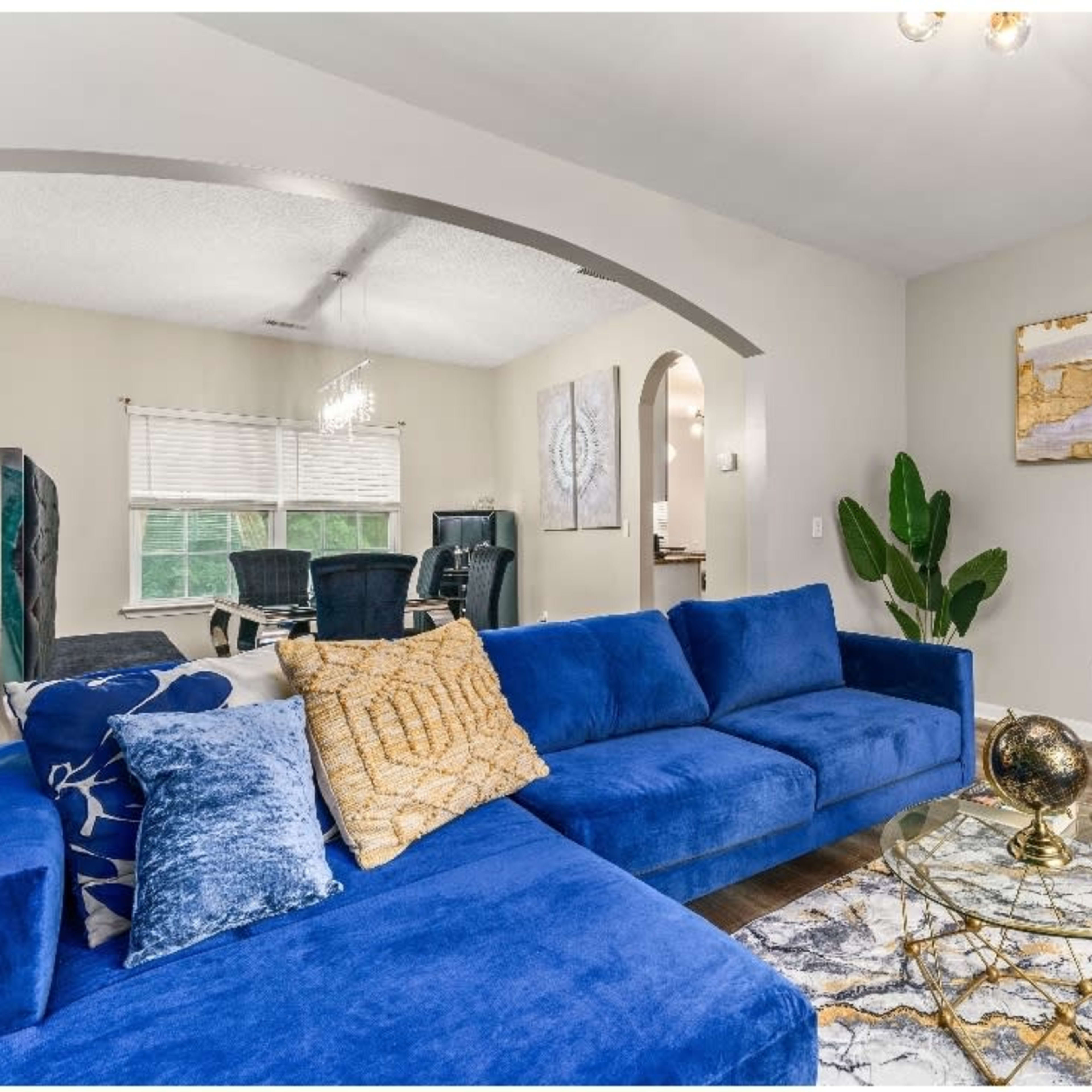 A blue sectional sofa faces a glass coffee table on a patterned rug, with a dining area visible in the background.