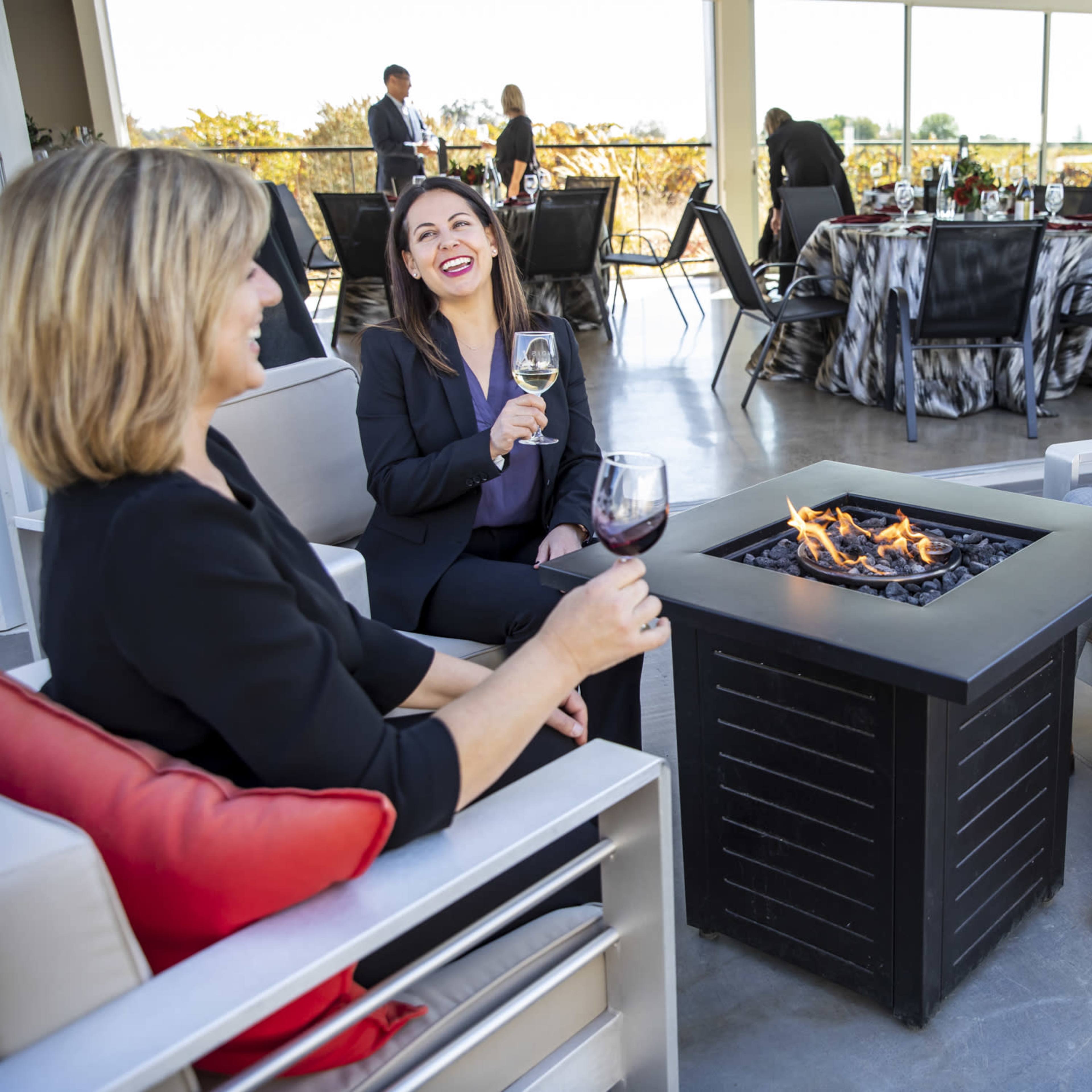 Three professionals sit around a fire pit, enjoying drinks in a modern indoor-outdoor event space with tables set for dining in the background.