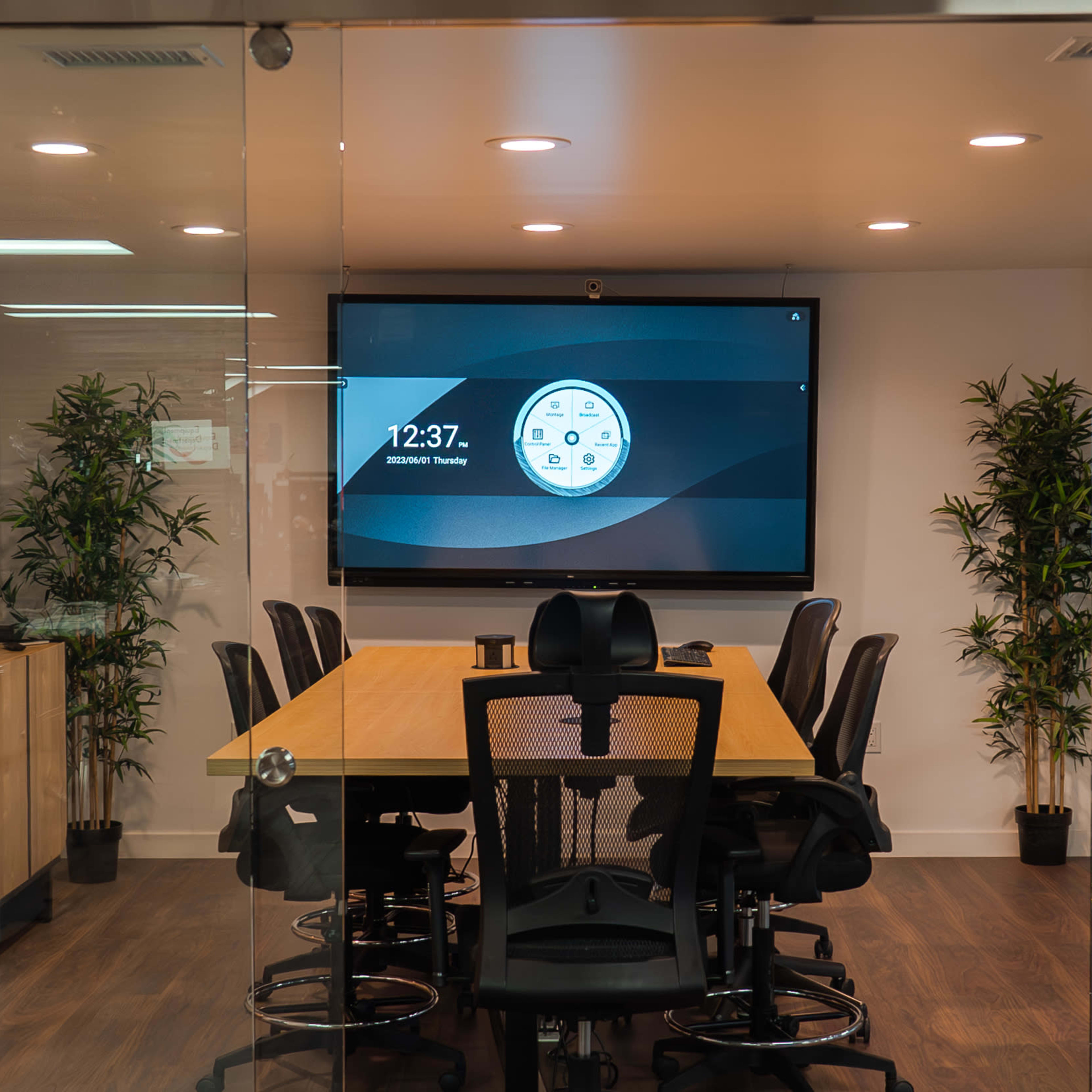 A modern conference room with a long wooden table, black mesh chairs, a large screen on the wall, and indoor plants.