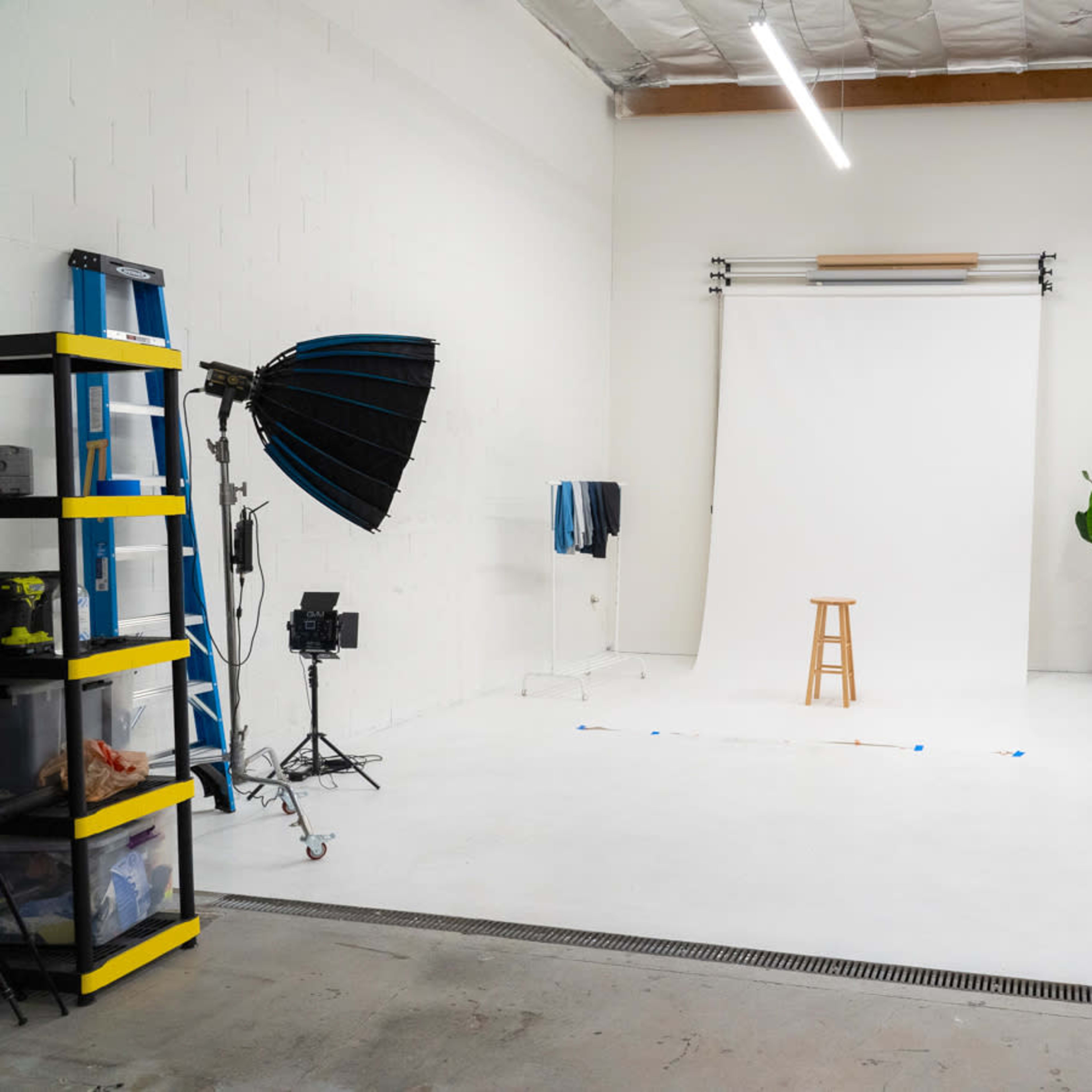 A photography studio with a white backdrop, a wooden stool in the center, two softbox lights on either side, and various equipment arranged around the space.