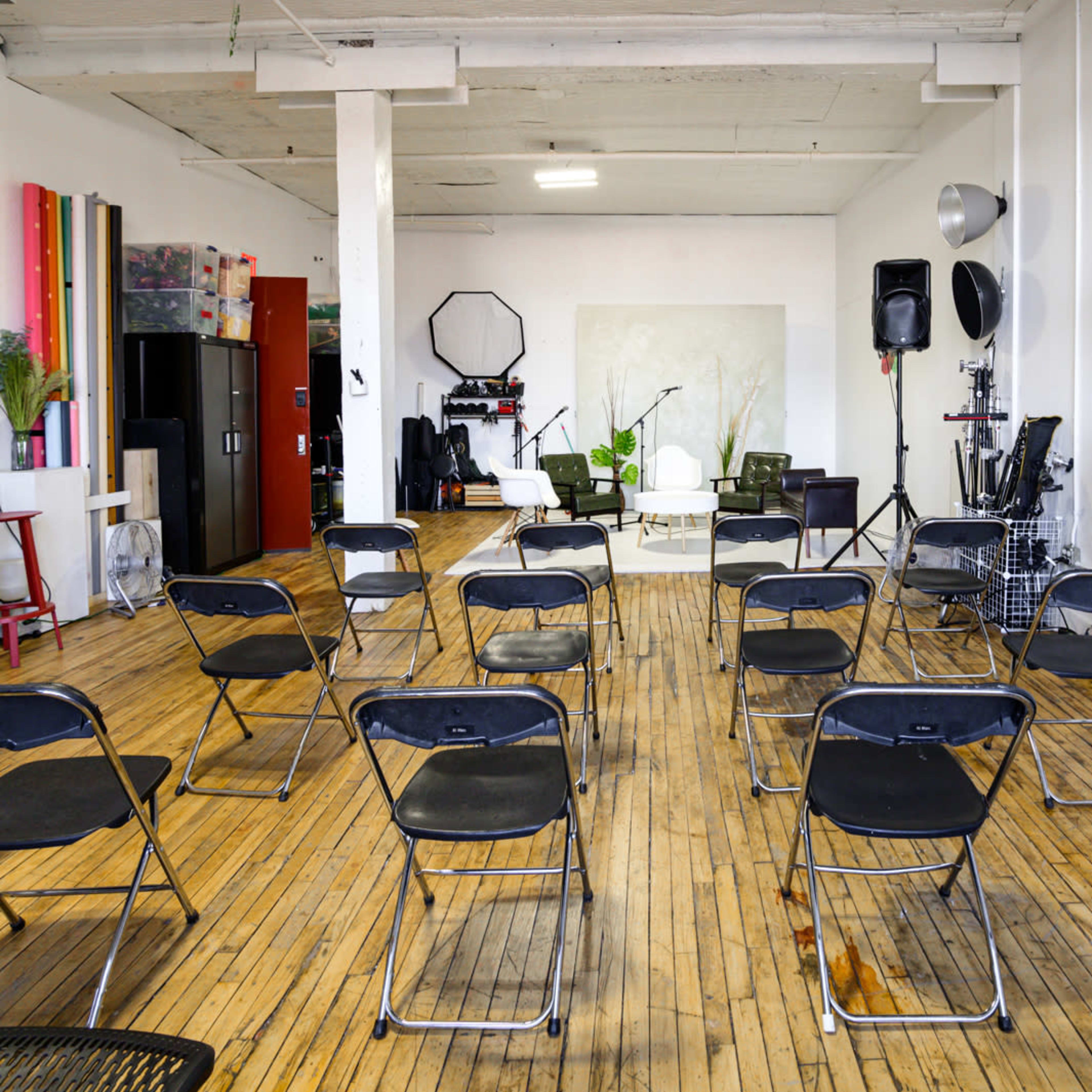 A spacious room features rows of black folding chairs arranged facing a blank wall with minimal decoration.