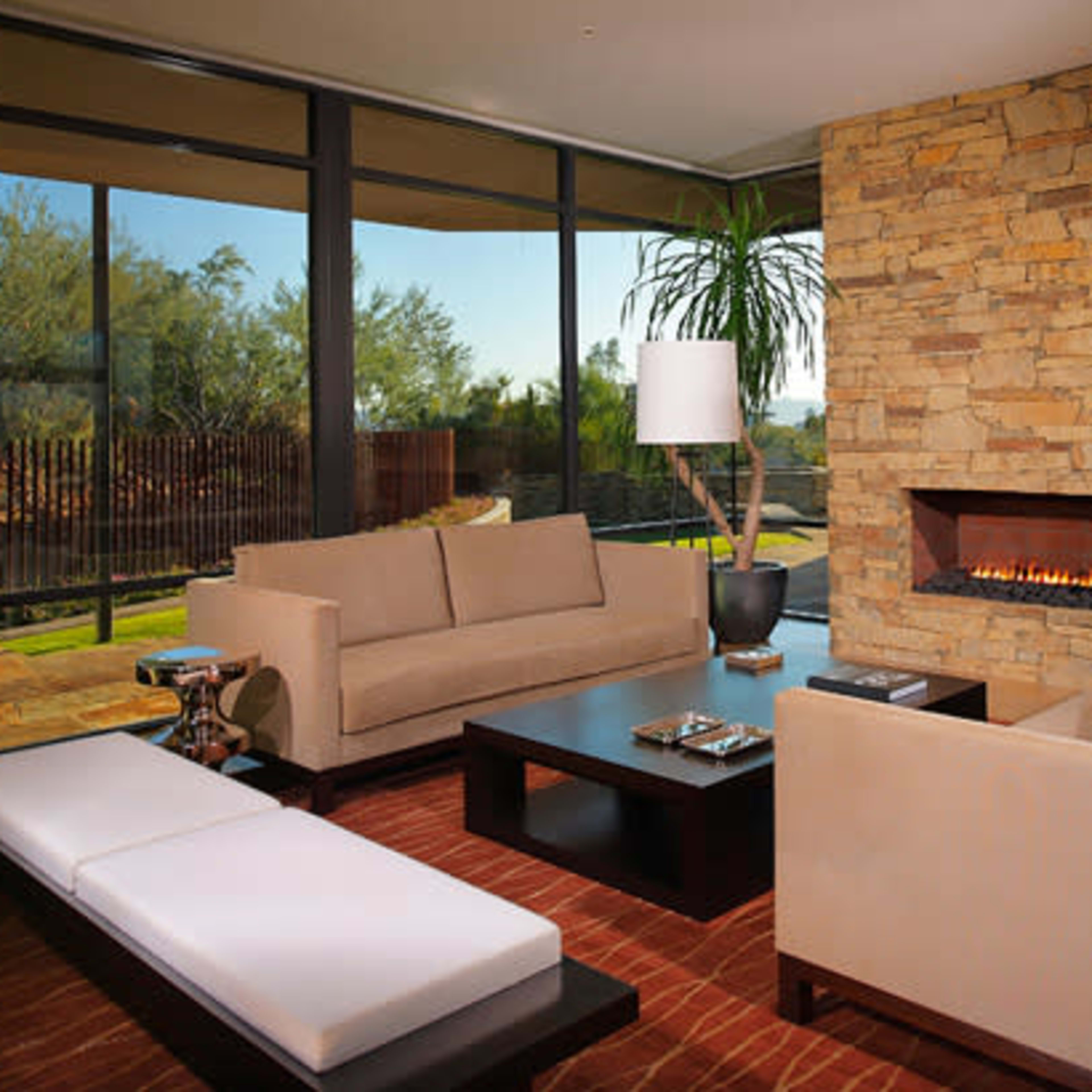 A modern living room features a stone fireplace, large windows, and contemporary furniture arranged around a coffee table.