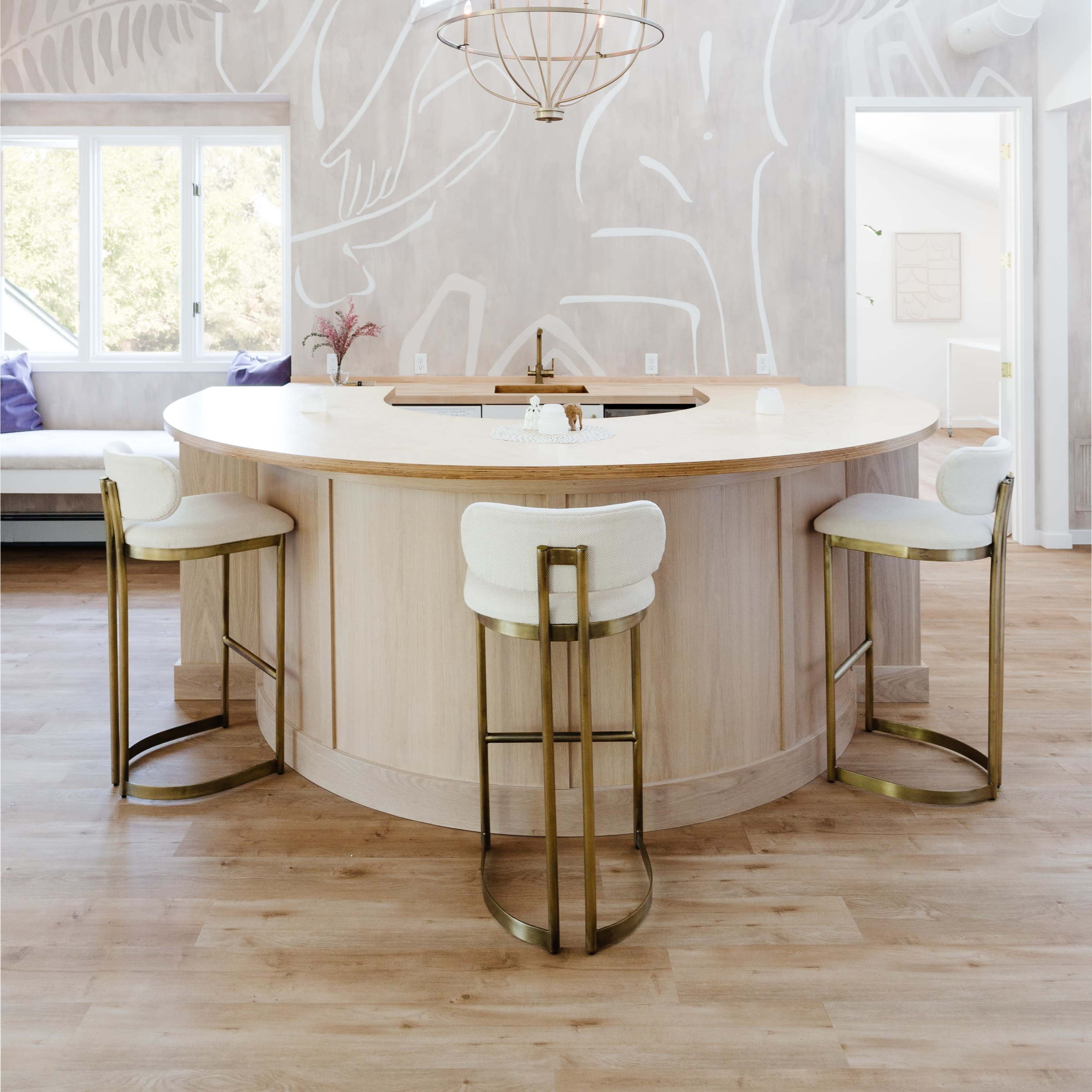A circular wooden kitchen island with four upholstered bar stools and a modern chandelier, set against a backdrop of large windows and a light-colored mural on the wall.