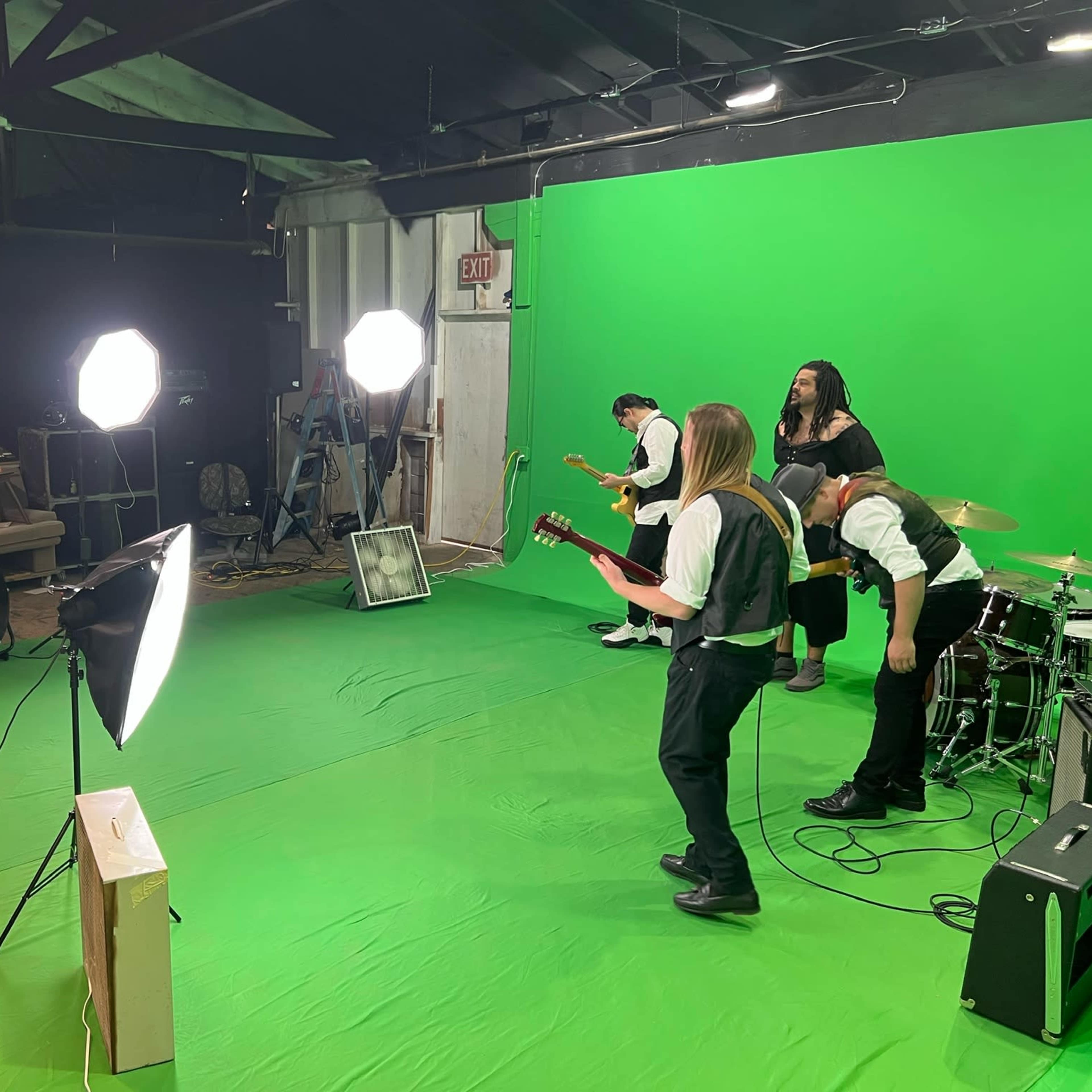 A band is performing on a green screen stage with two bright lights and a camera recording from the side.