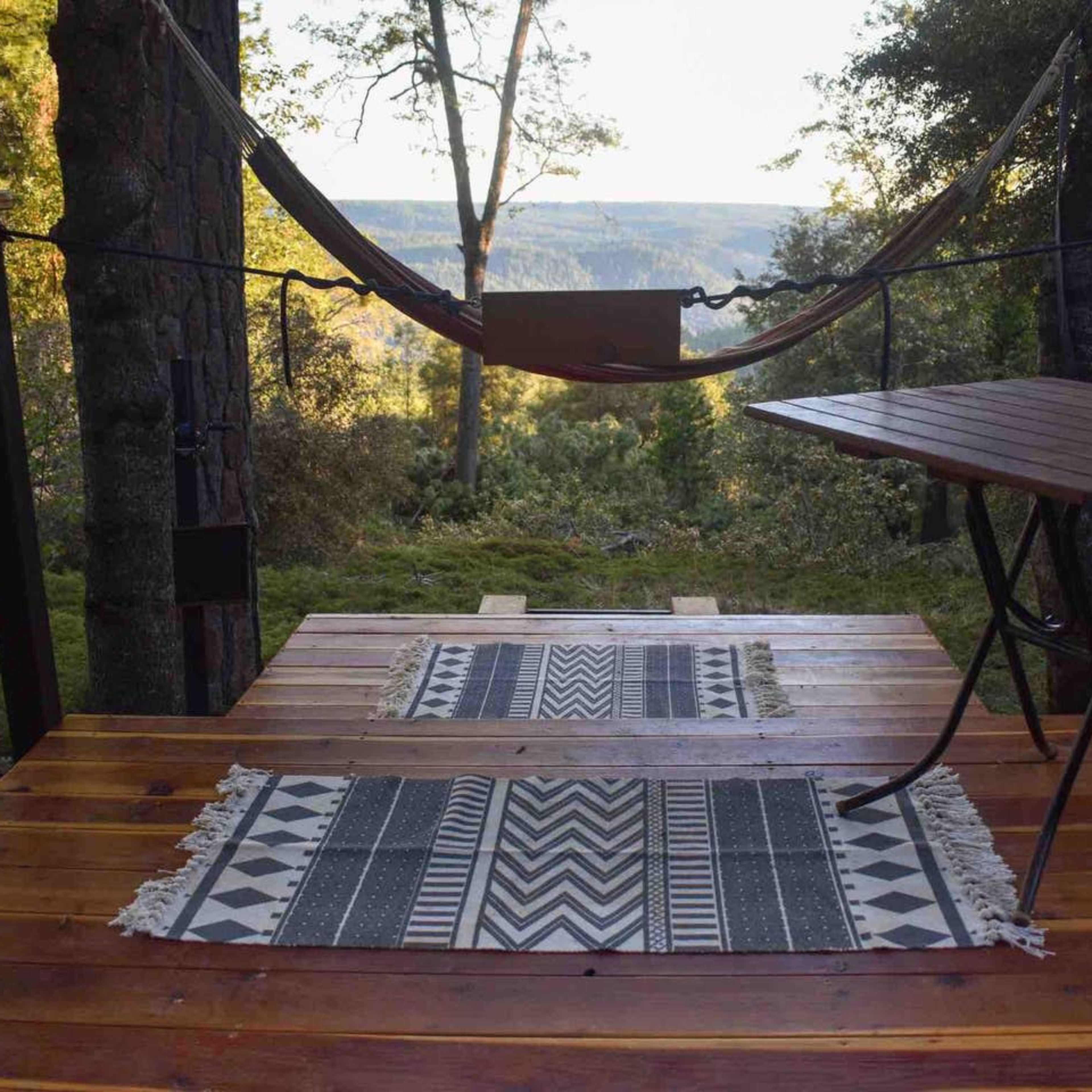 A wooden deck with a hammock suspended between two trees, accompanied by a small table and patterned rugs, overlooking a scenic view of hills and trees.