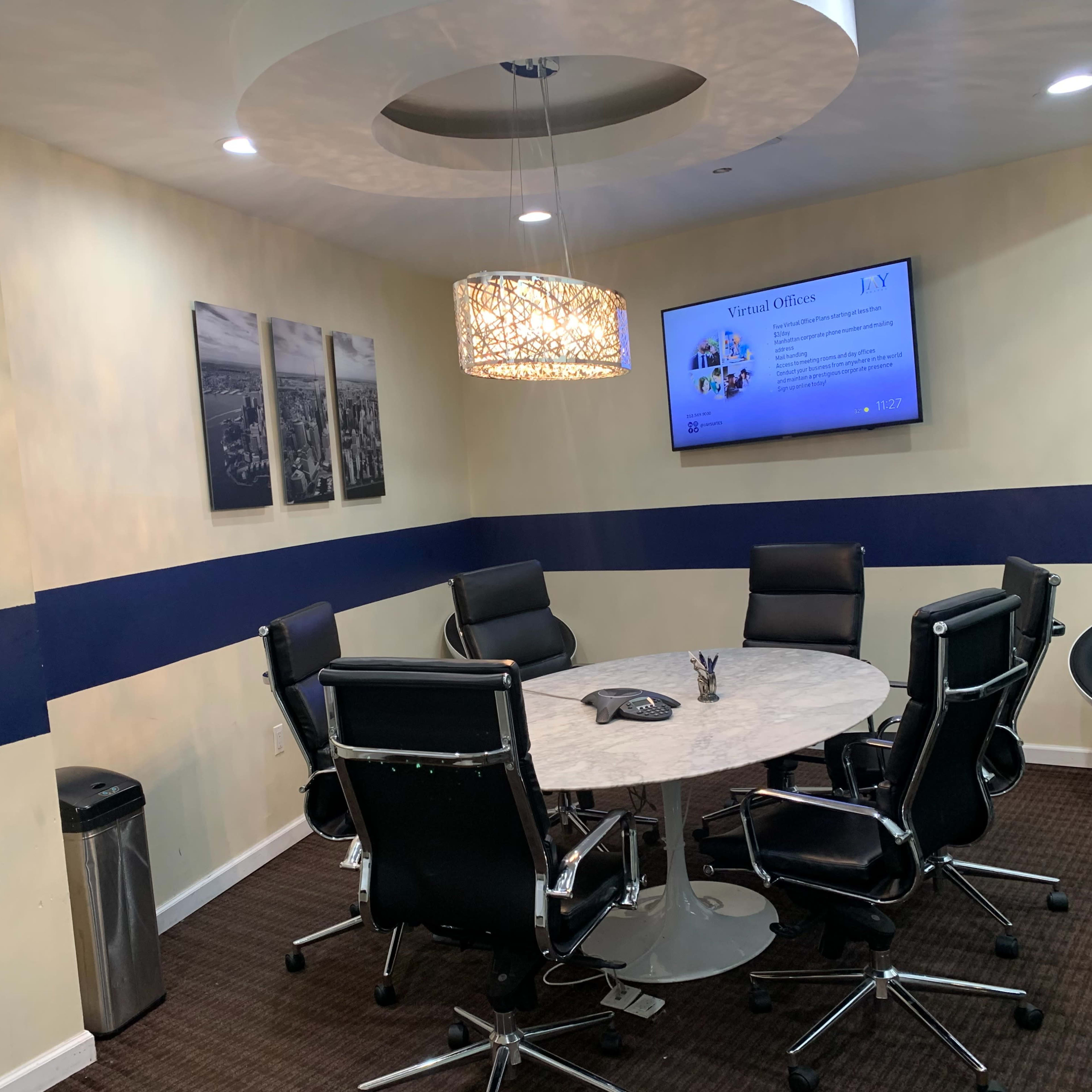 A conference room with a round table, six black chairs, a wall-mounted screen displaying information, and three framed photos on the wall.