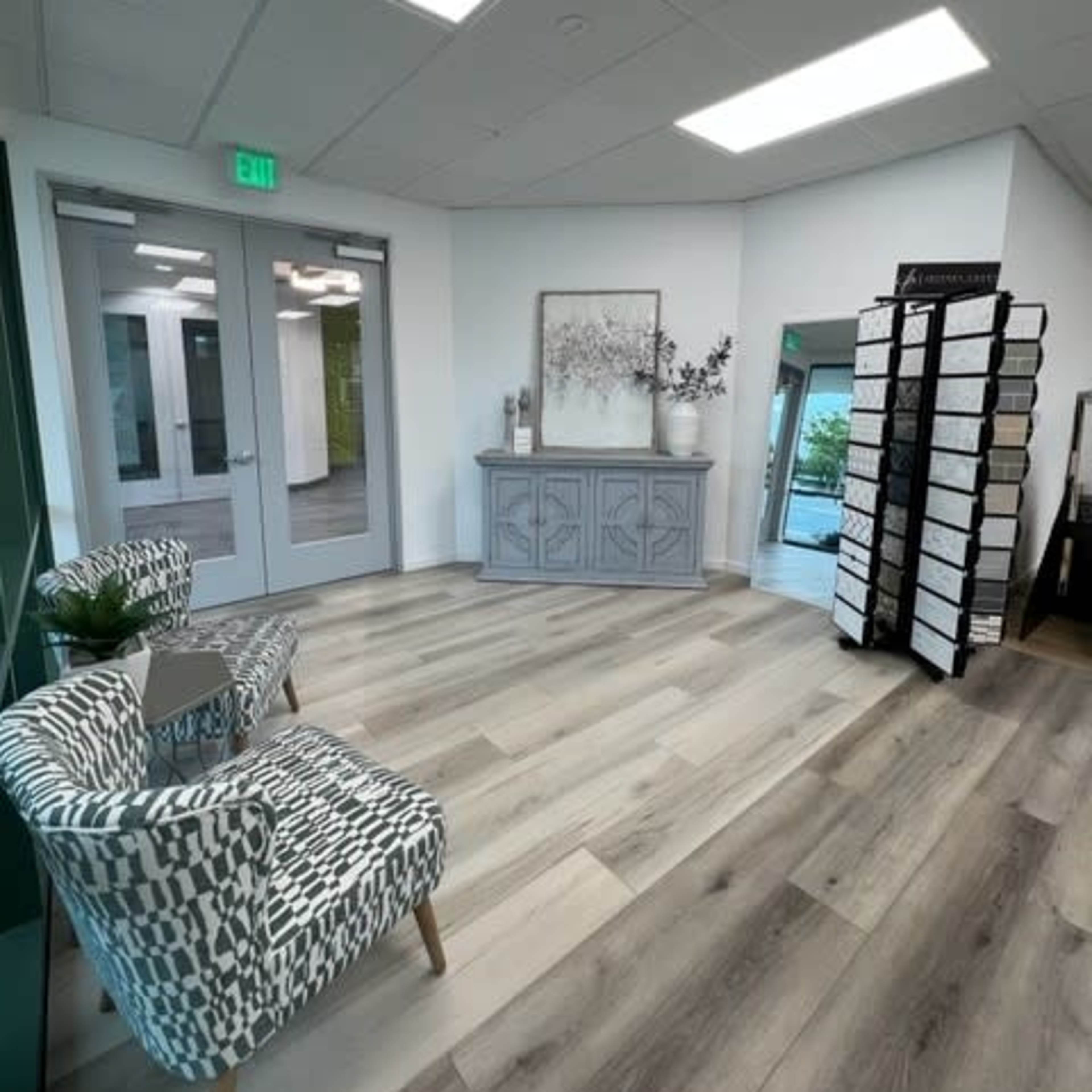 A spacious interior room with a light wood floor, a decorative gray cabinet, two patterned chairs, and a display rack filled with samples.