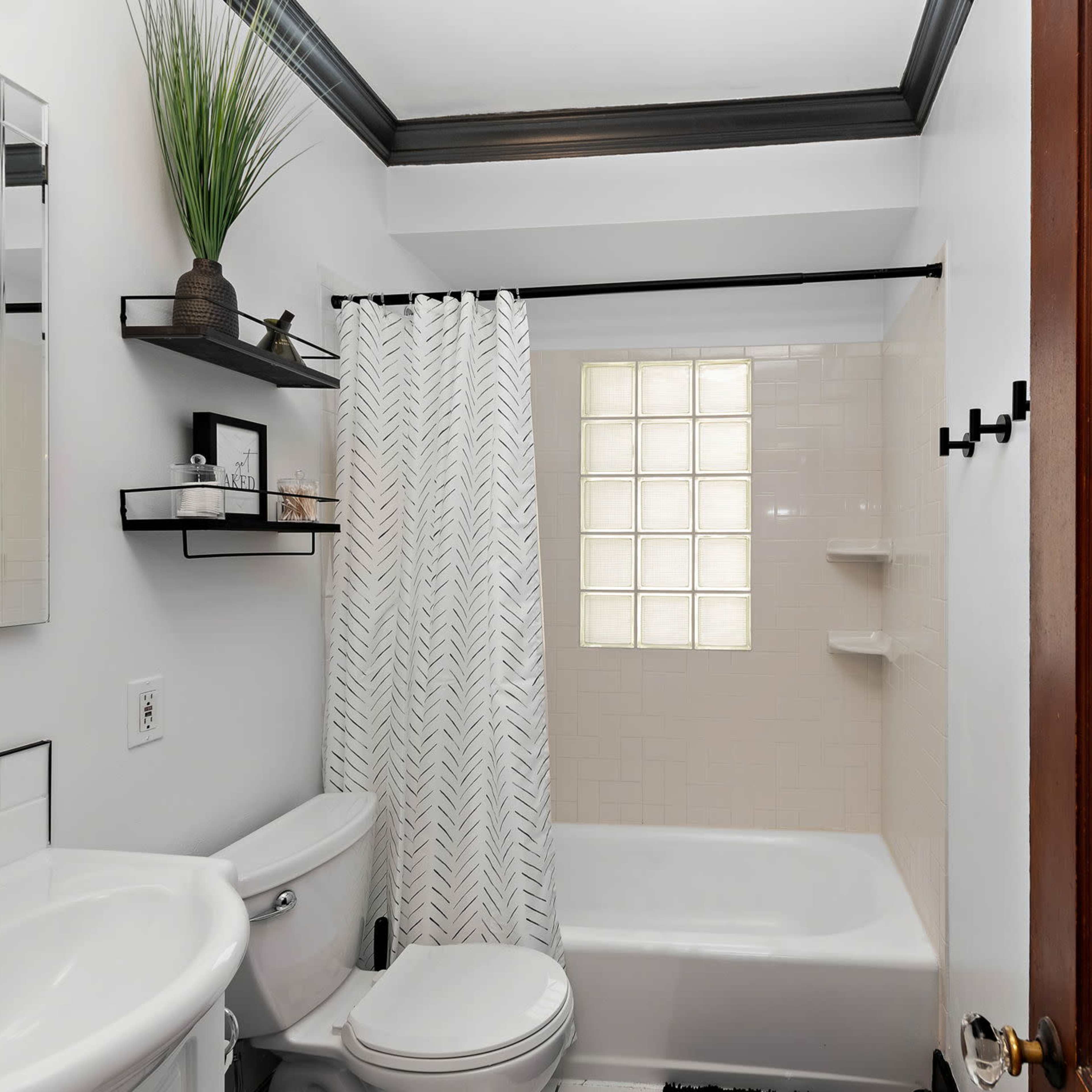 A small bathroom featuring a toilet, sink, and a bathtub with a shower, complemented by a window made of glass blocks and minimal shelf storage.