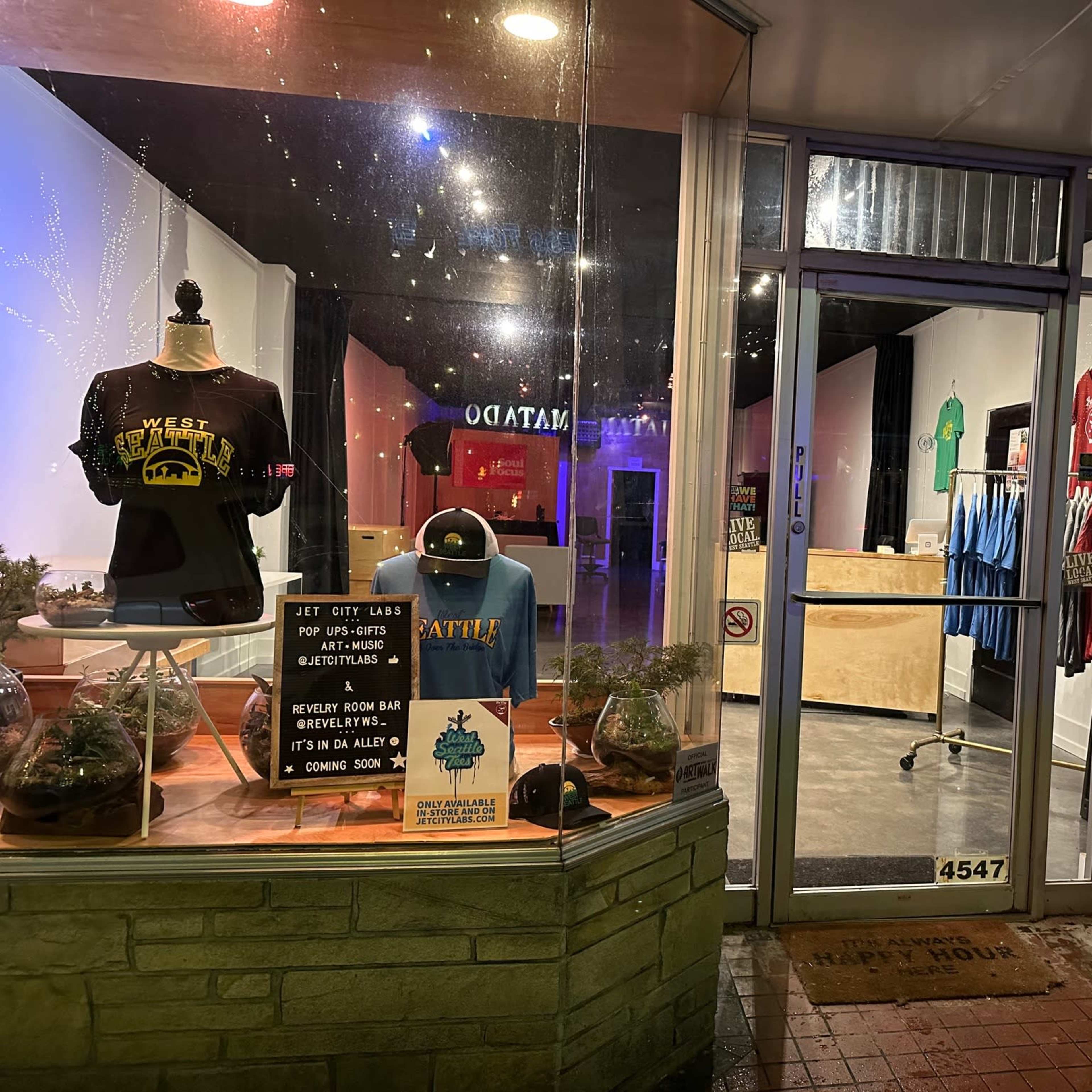 The storefront displays t-shirts and promotional signs in a well-lit window, showcasing a variety of merchandise and upcoming events.
