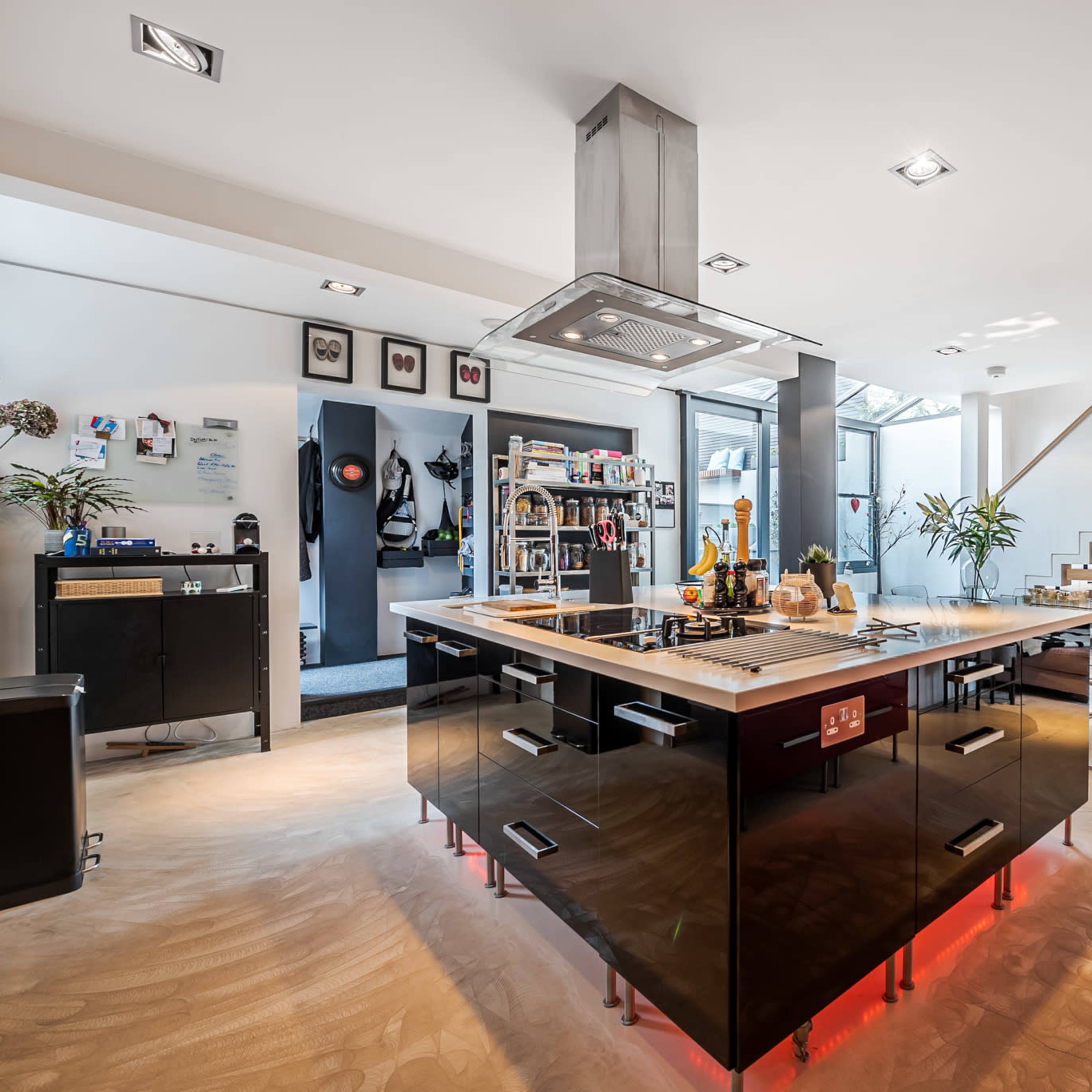 A modern kitchen features a central island with a stovetop and pendant lighting, surrounded by sleek cabinetry and an open living area with a staircase.